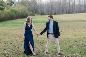 Couple in formal attire holding hands while walking through grassy field.
