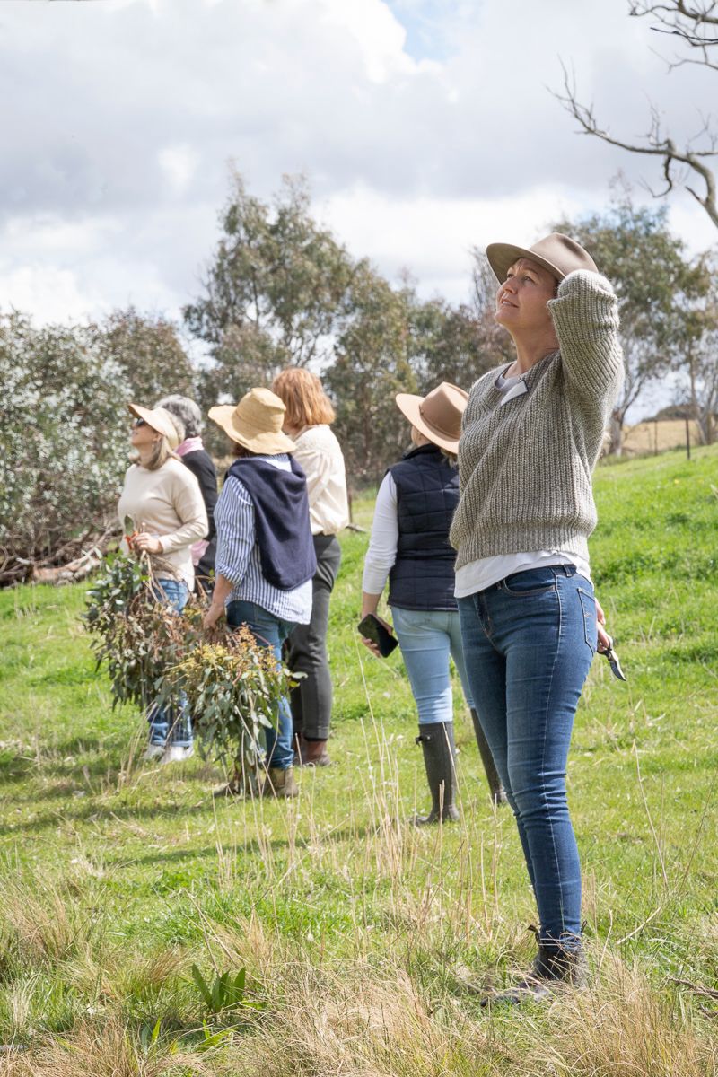 A line of women standing in a paddock wearing hats holding foraged gum leaves. Event photography by Em Callaghan Photography.