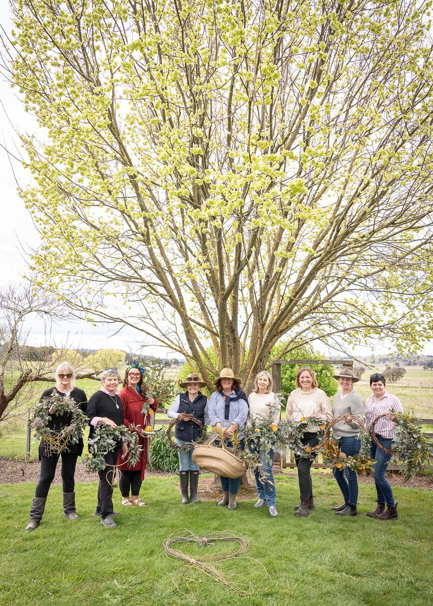 A group of women standing under a tree holding handmade wreaths in a country garden. Event photography by Em Callaghan Photography.