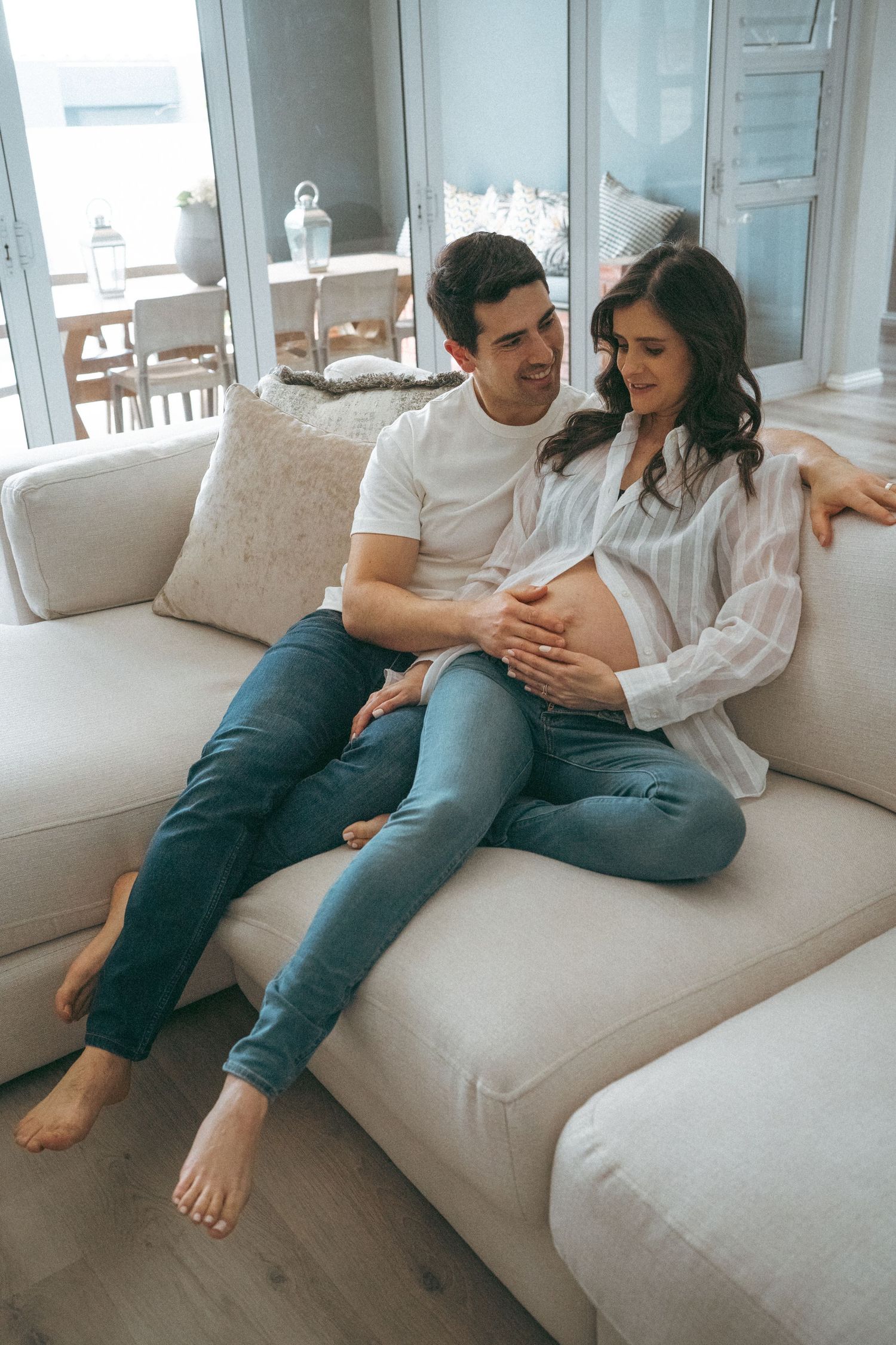 Expectant couple sharing an intimate moment while relaxing on a beige couch in a bright living room.