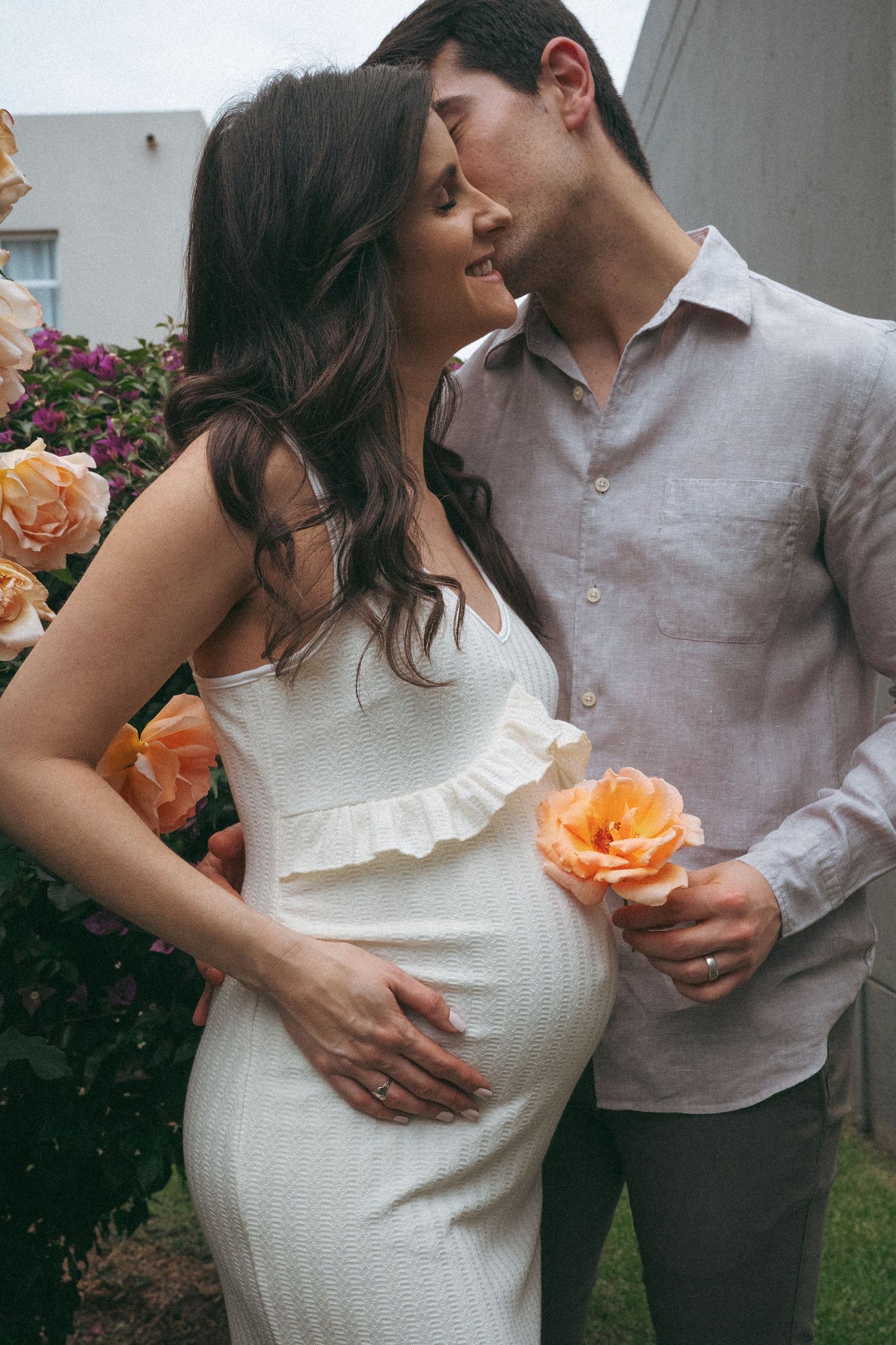 Romantic maternity photo with couple by peach rose bush, woman in white dress.
