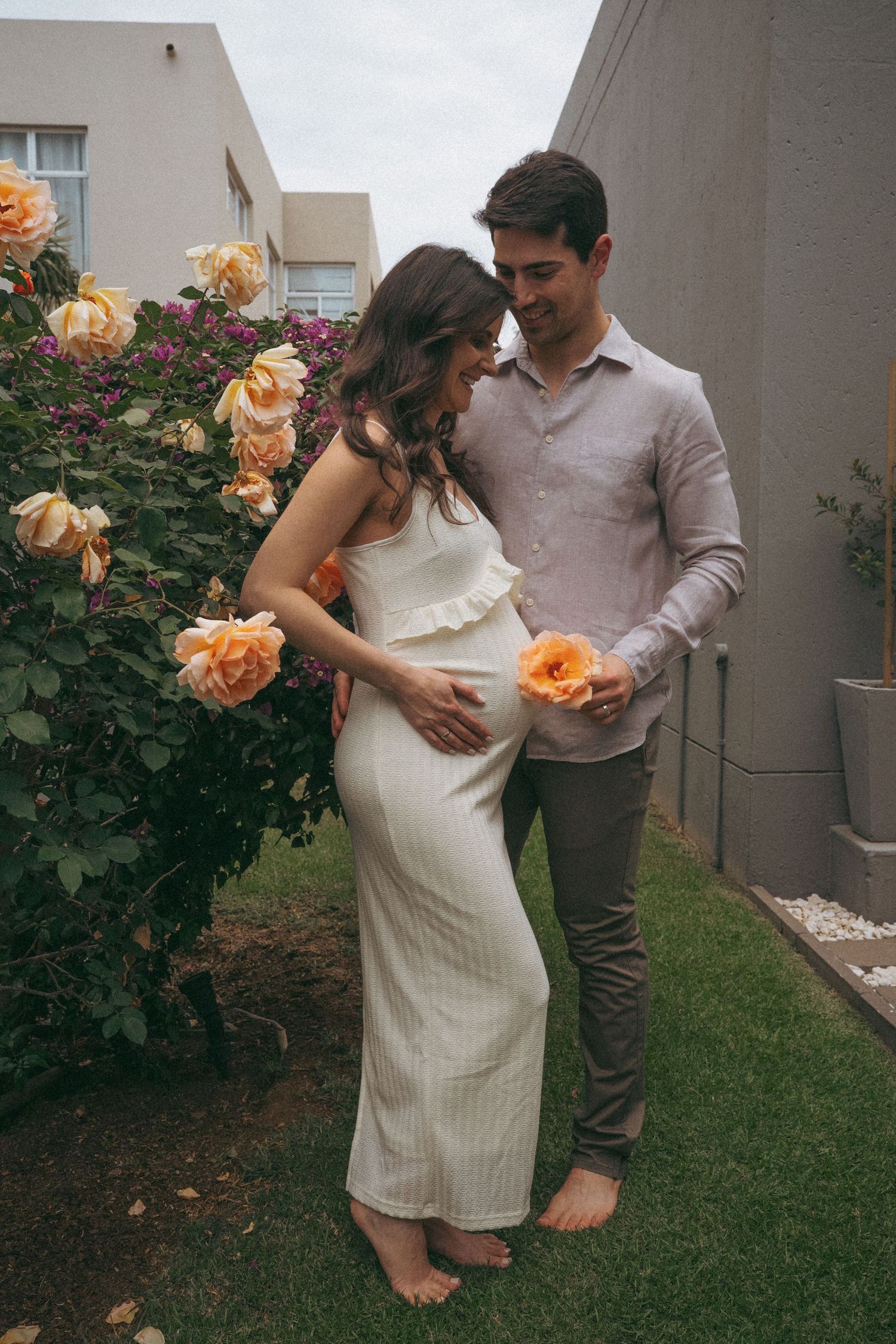 Couple poses by blooming peach roses in garden during maternity photoshoot.