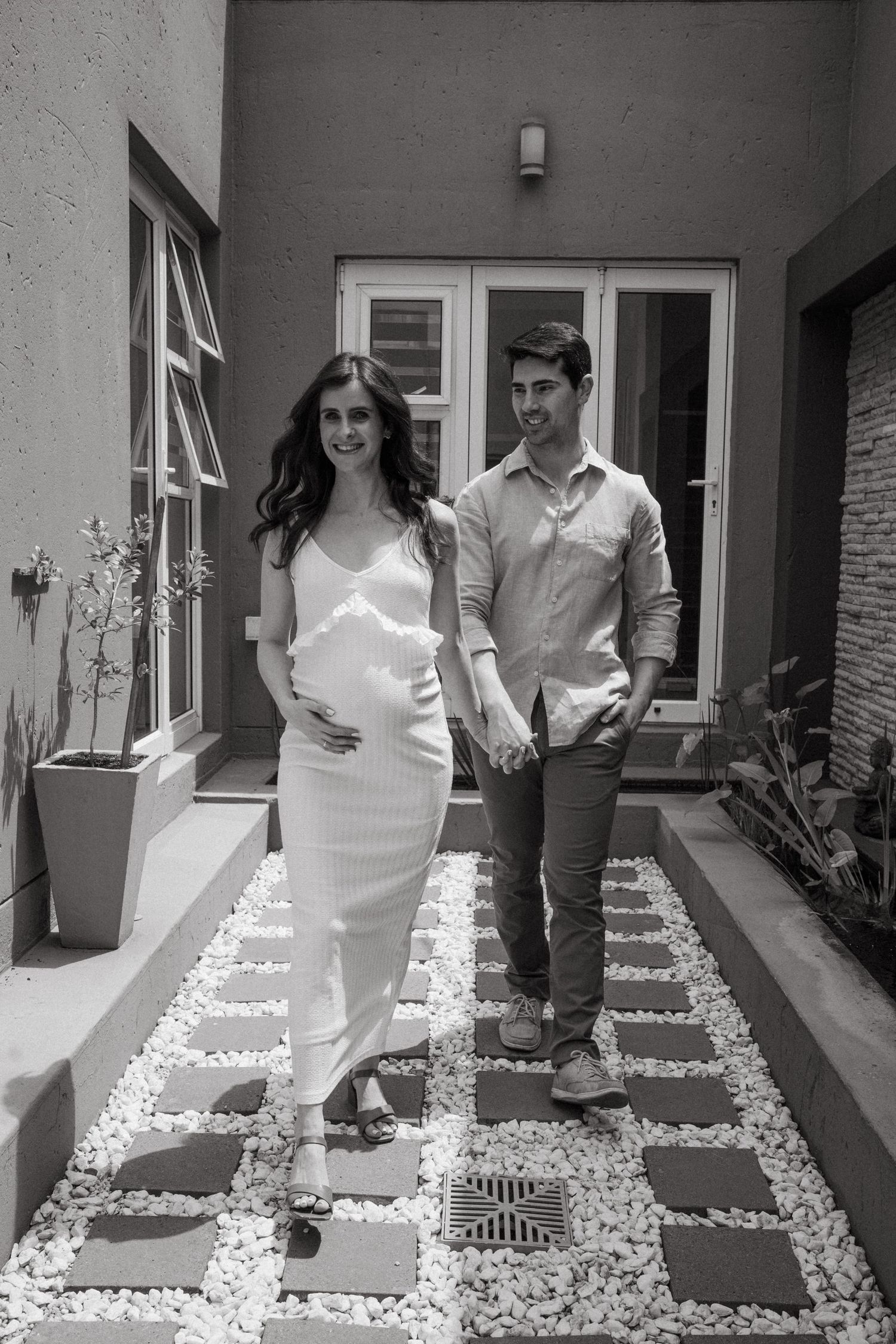 Couple walking hand in hand on stone pathway outside their home in black and white photo.