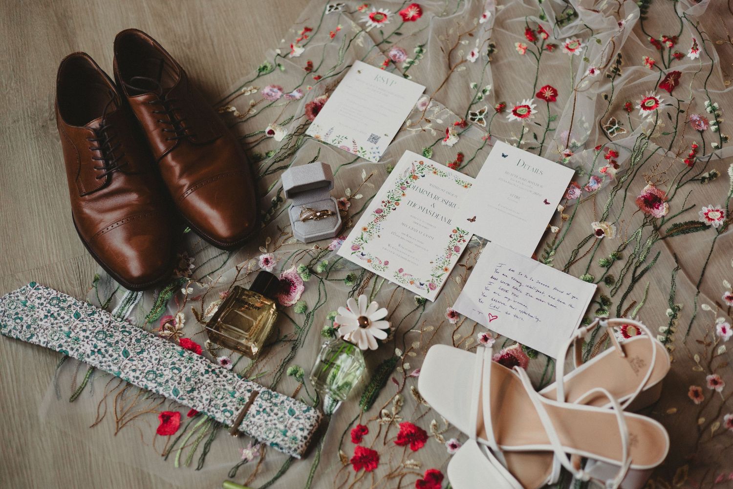 Wedding details flatlay with brown leather shoes, invitations, and pink heels on a floral fabric.