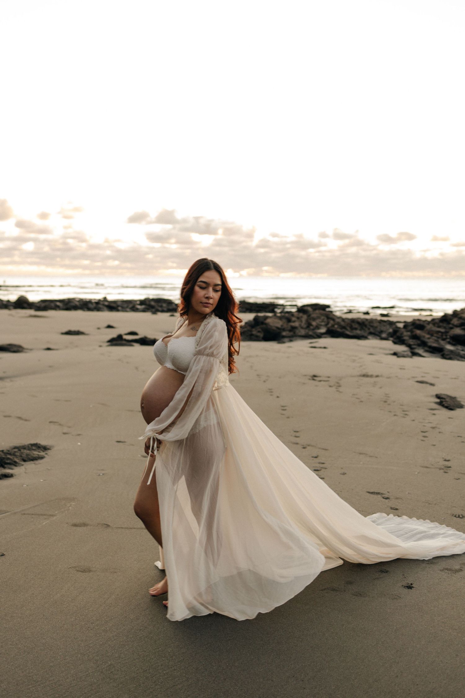 Romantic beach maternity photoshoot featuring flowing white dress at golden hour.