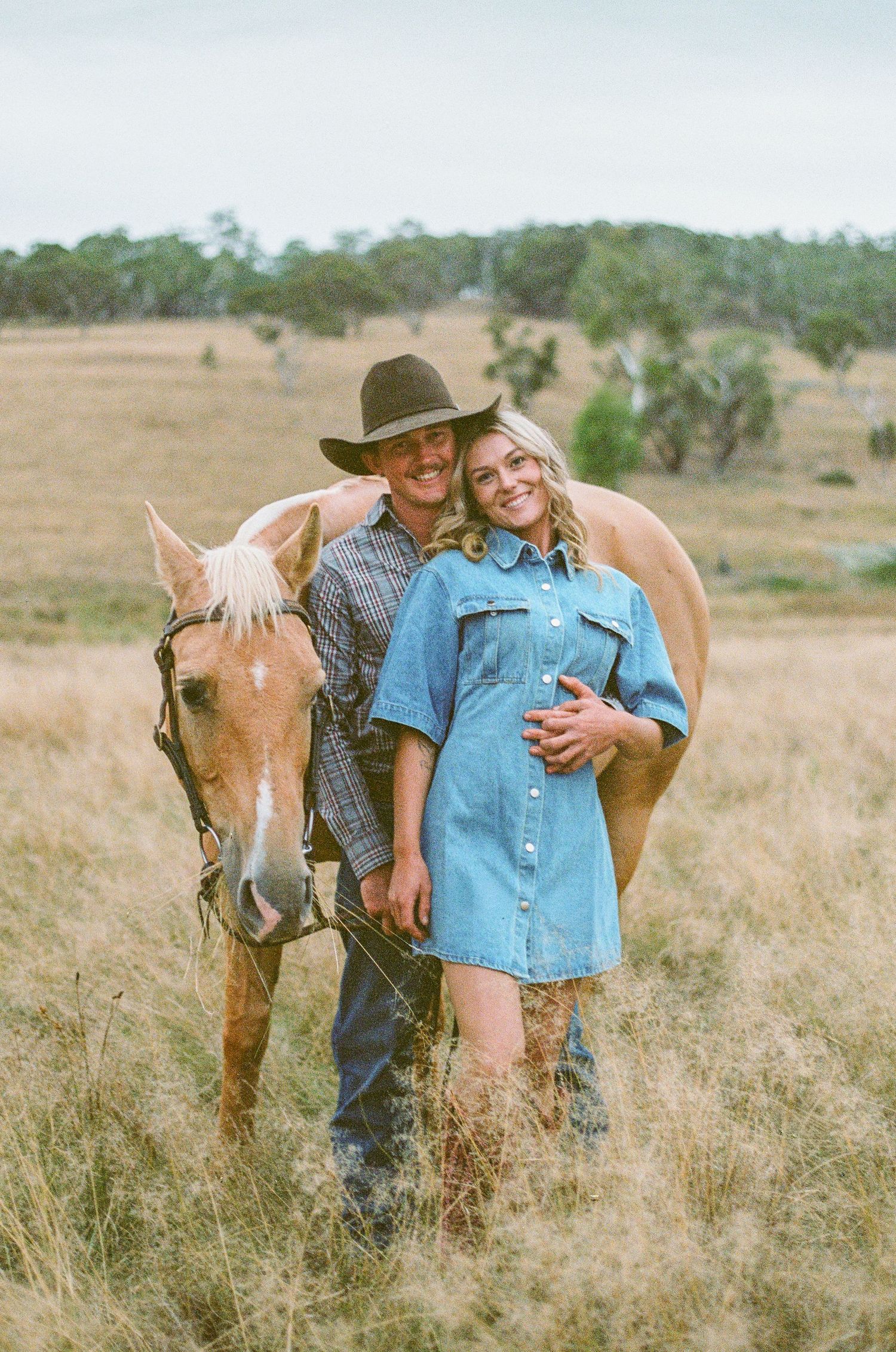 man in cowboy hat hugs girl in denim dress and cowgirl boots during engagement photoshoot while holding horse