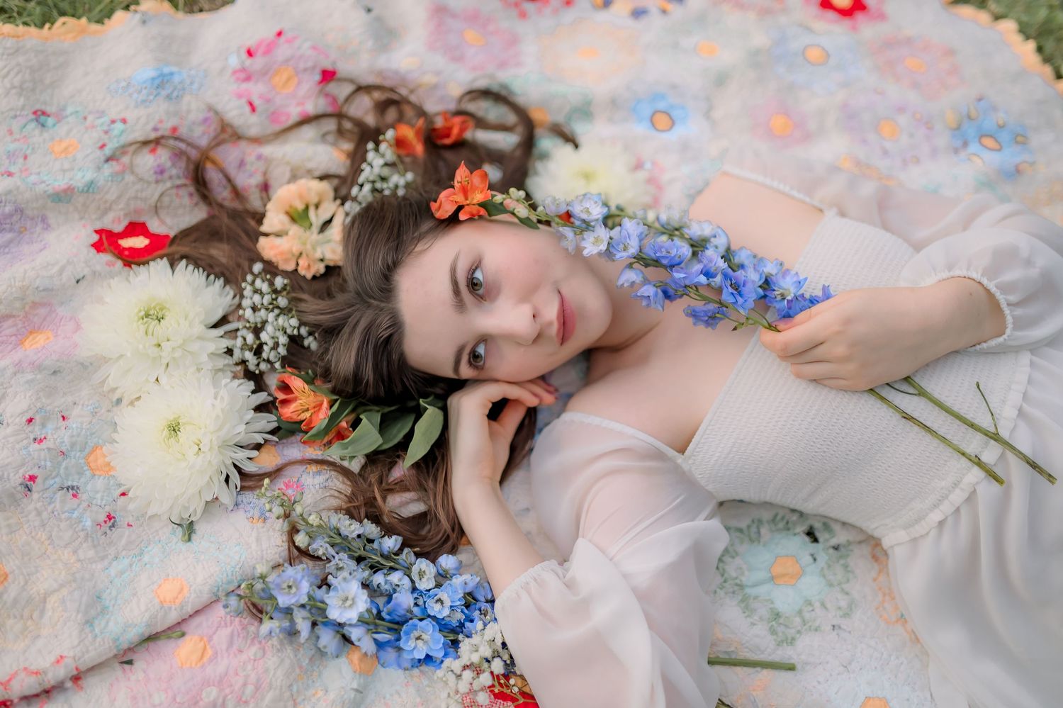 Ethereal portrait on floral blanket with scattered delphinium flowers and a flower crown surrounded by soft pastel colors.