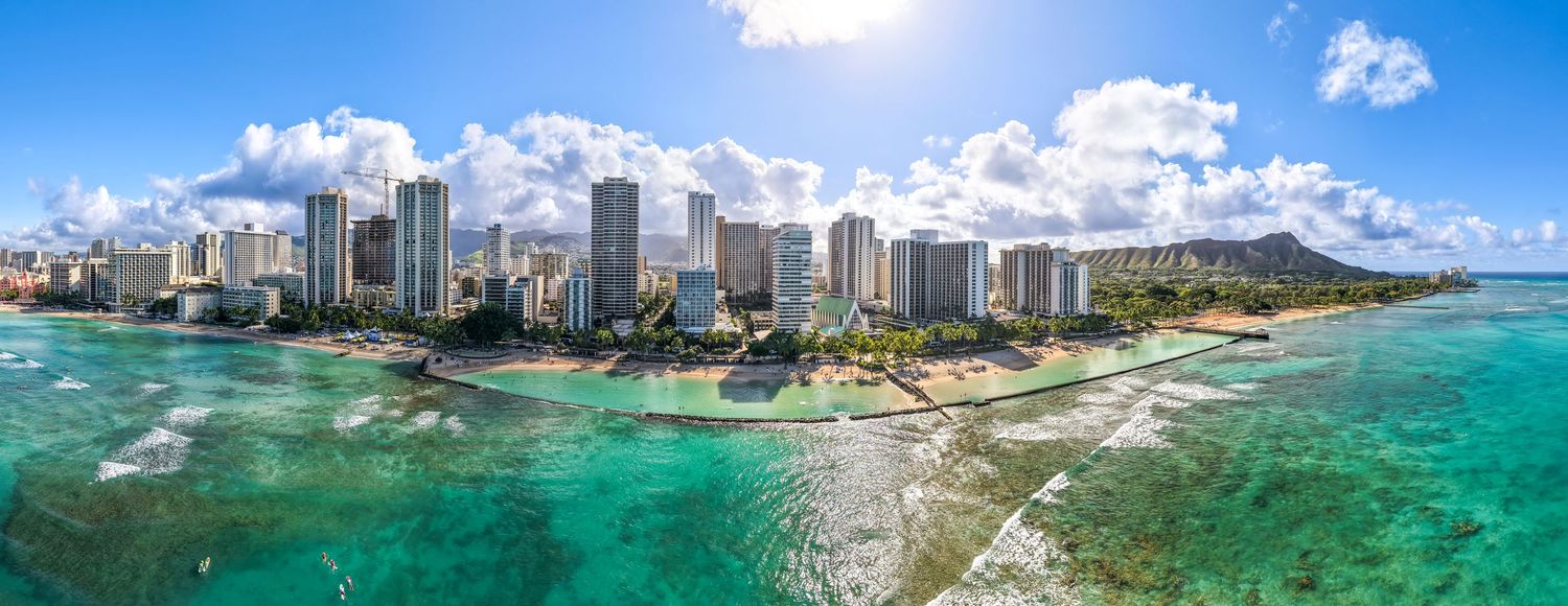 Aerial panorama of Waikiki Beach showing high-rise hotels, turquoise waters, and coastal landscape.