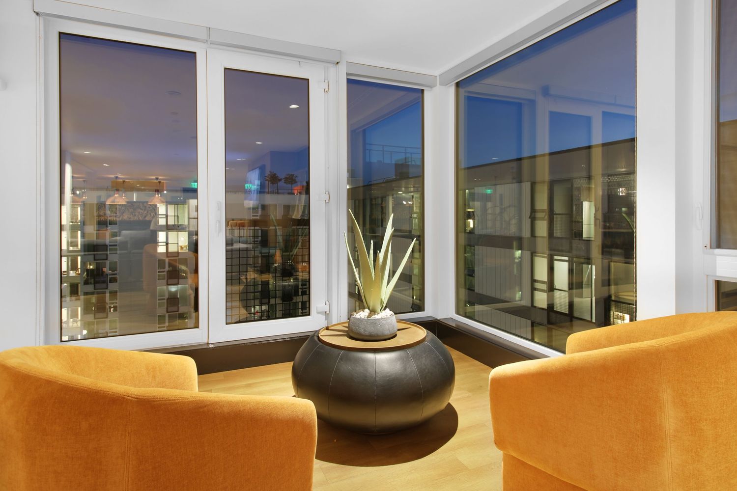 Modern corner apartment with floor-to-ceiling windows, orange accent chairs and a potted plant on the window ledge at dusk.
