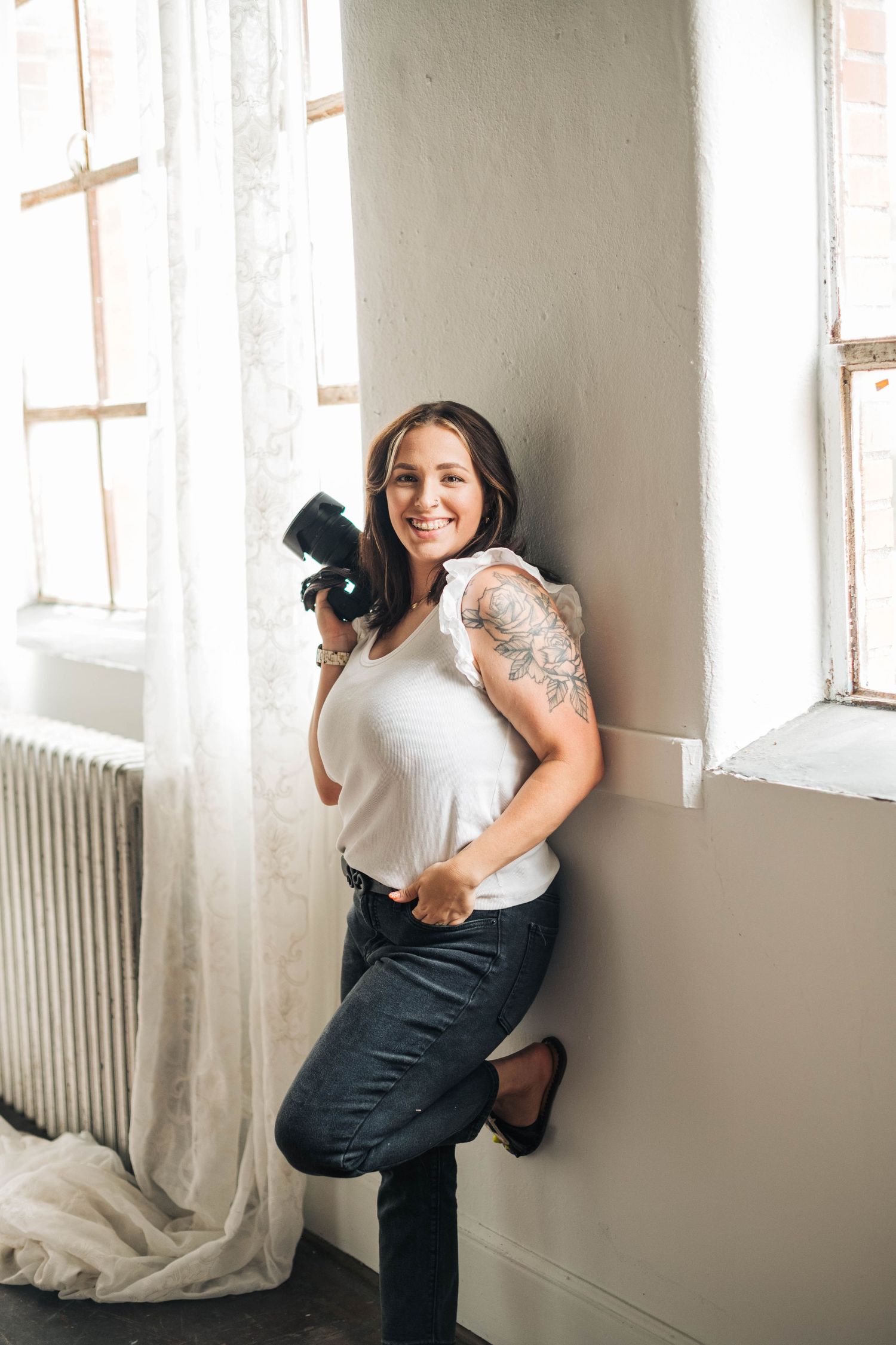 A photographer holds a camera while posing against a white wall near a window.