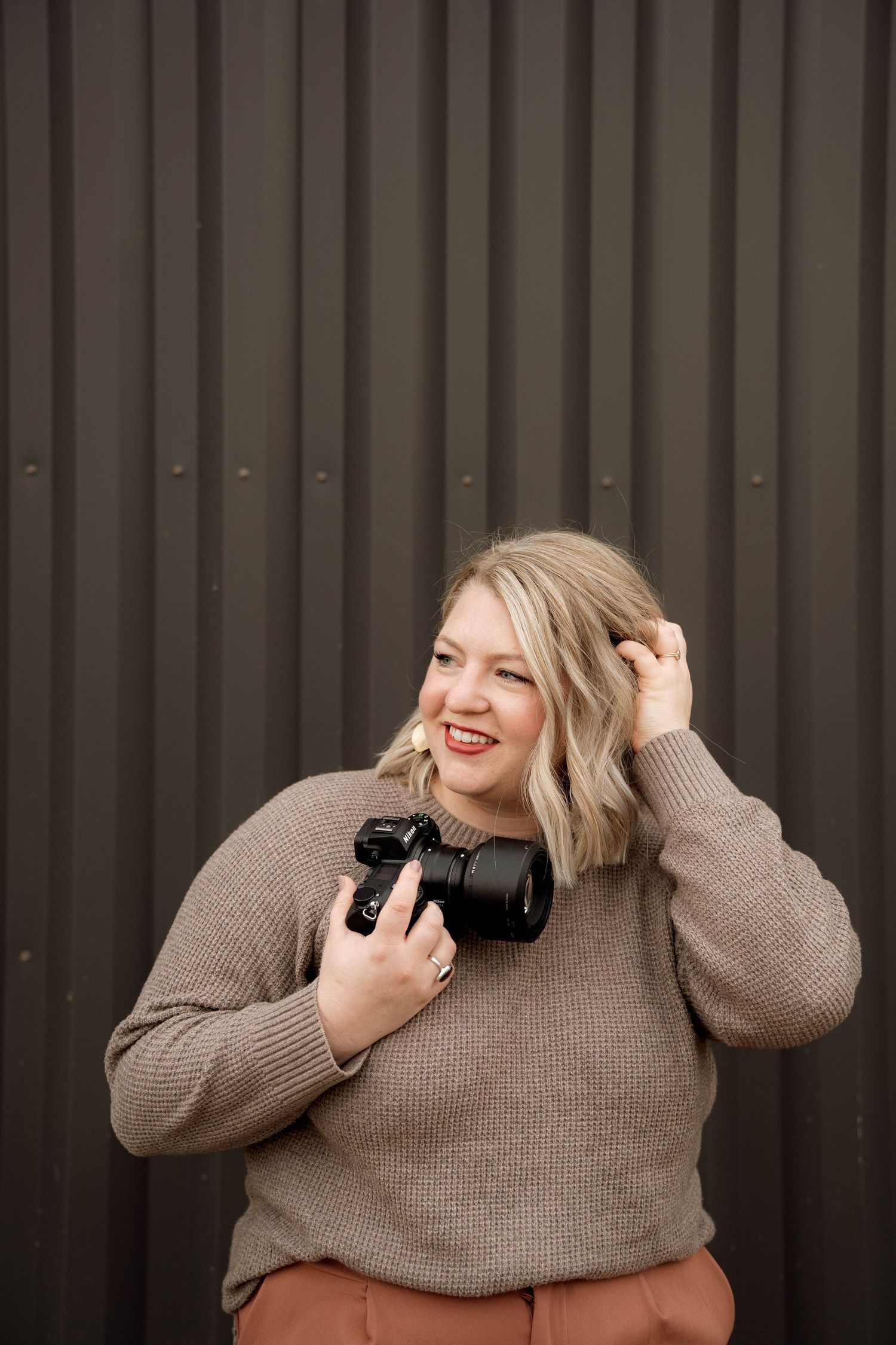 Professional photographer in tan sweater holding camera against dark wall.