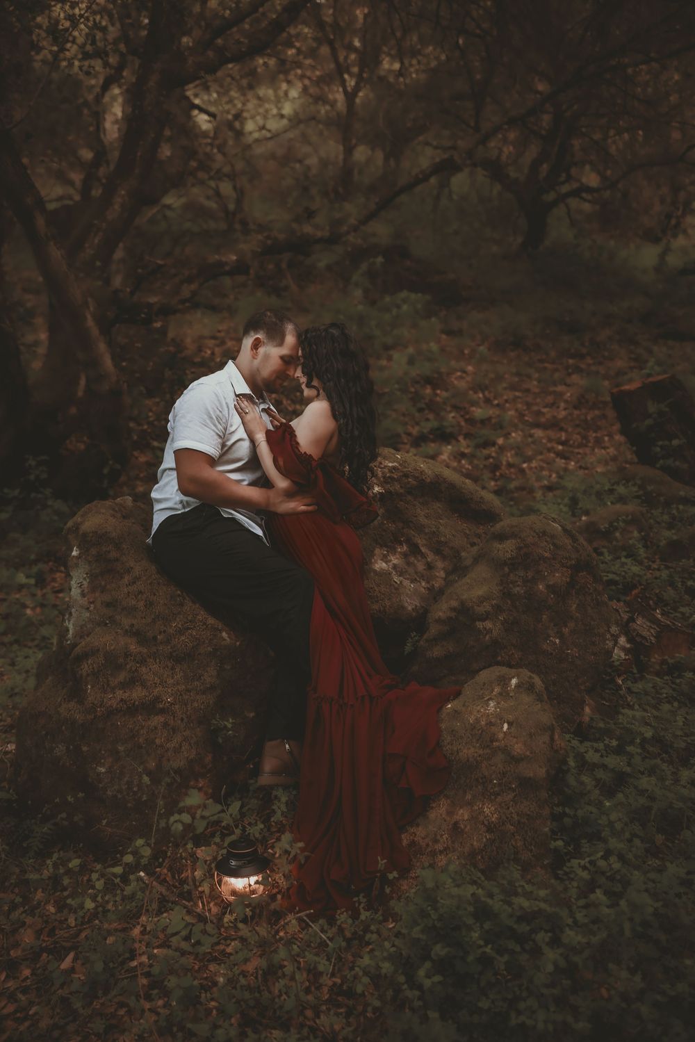 Couple sharing a romantic kiss on rocks in a dark forest, woman in flowing red dress with lantern.