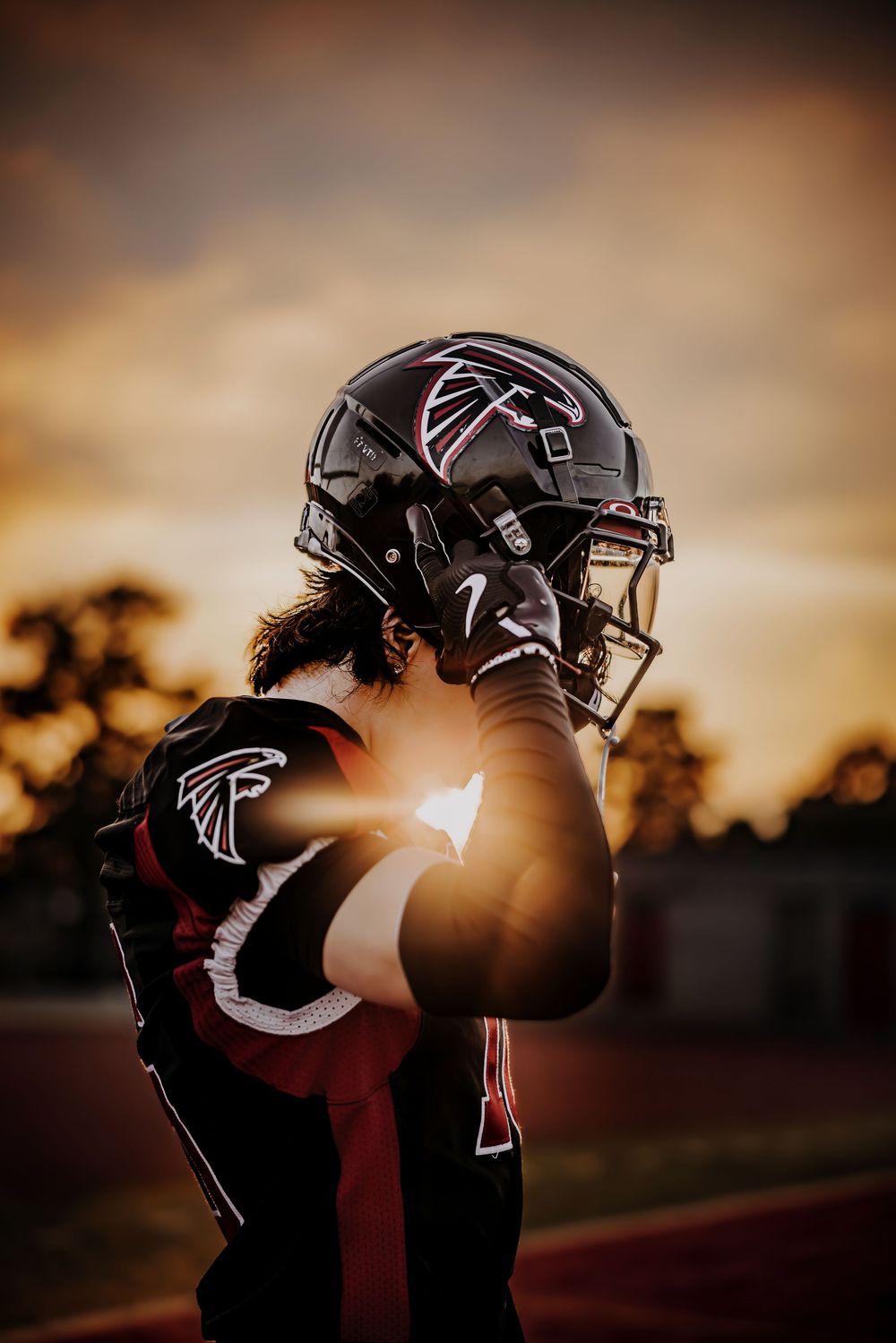 Silhouetted football player in Atlanta Falcons uniform holds helmet against dramatic sunset sky during practice.