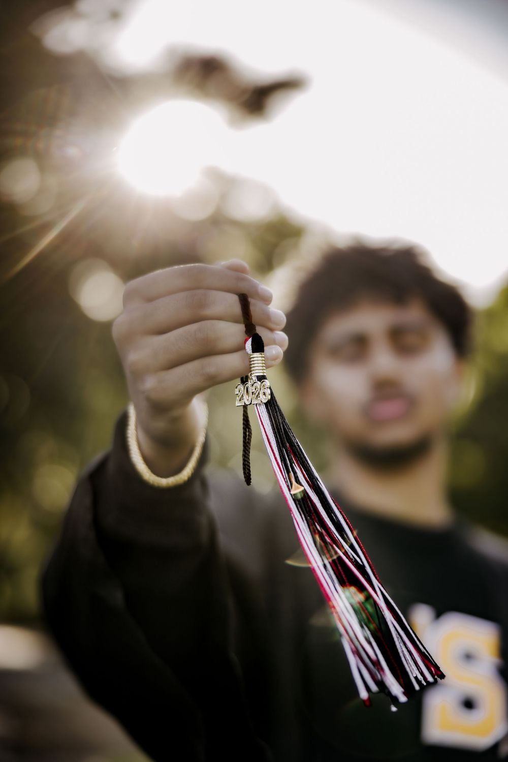A young man holds up a colorful tassel keychain toward the camera with sunlight glowing behind him outdoors.