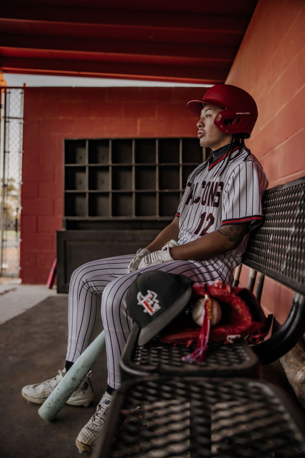 Baseball player in Falcons pinstripe uniform sits in dugout wearing red helmet, holding bat and glove.