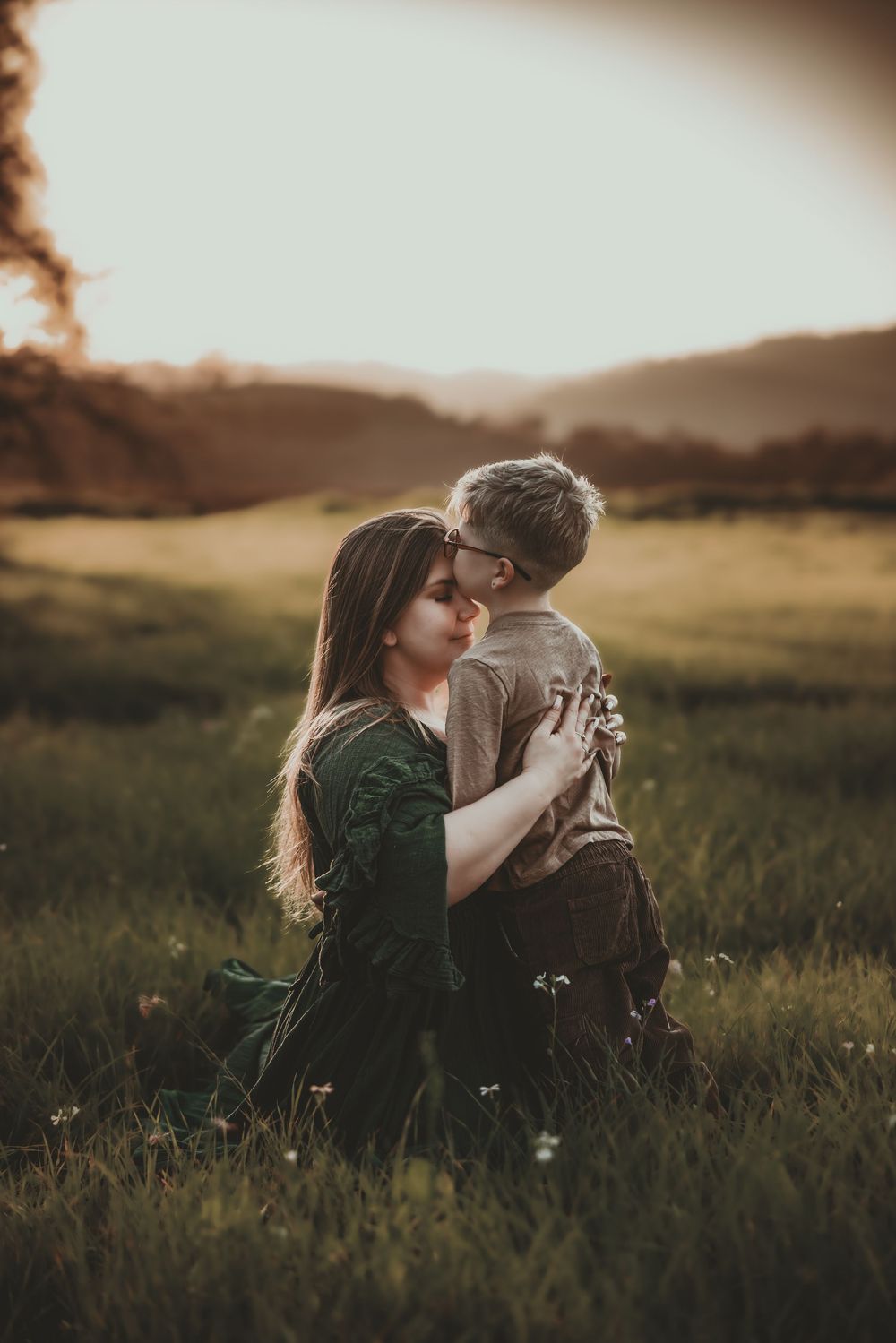 A mother and young son share a tender nose-to-nose moment in a sunlit green meadow at golden hour.