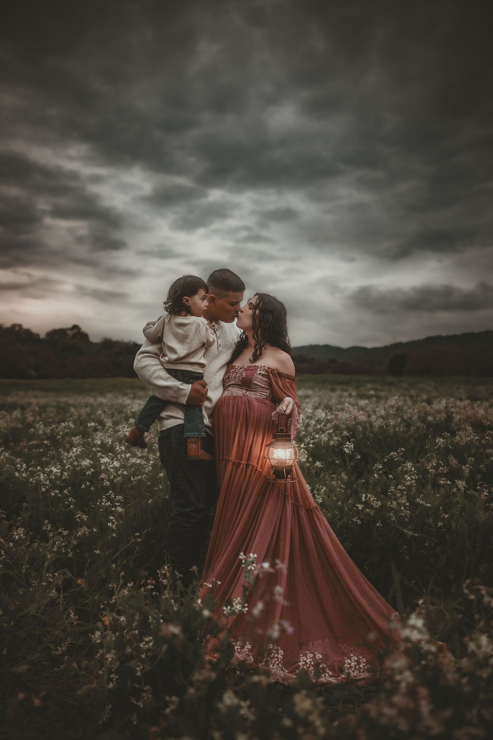 A couple in vintage-style clothing embraces romantically in a moody wildflower field under dramatic stormy skies.
