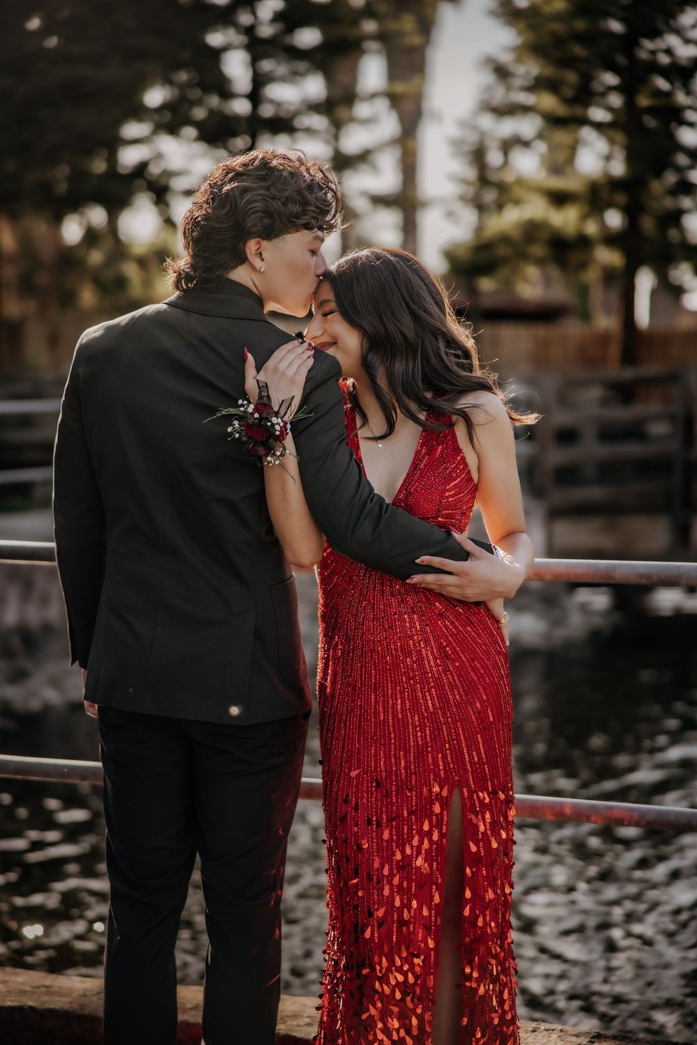 A couple in prom attire embraces near a railing, the girl in a red sequin gown and the boy in a black suit.