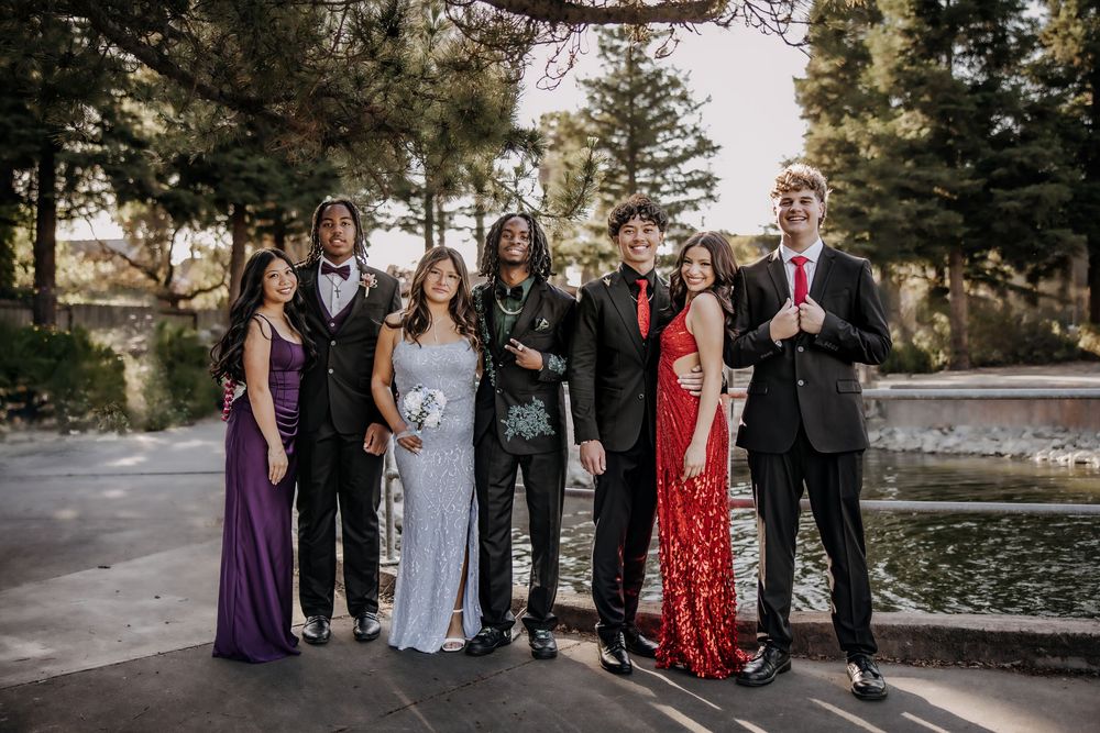 Group of seven teens dressed in formal prom attire posing outdoors near a fountain with trees in background.