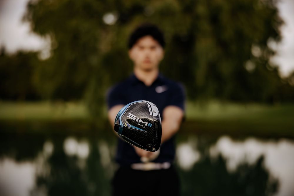 Golfer holding a Ping golf club driver toward the camera near a reflective pond with green trees in the background.