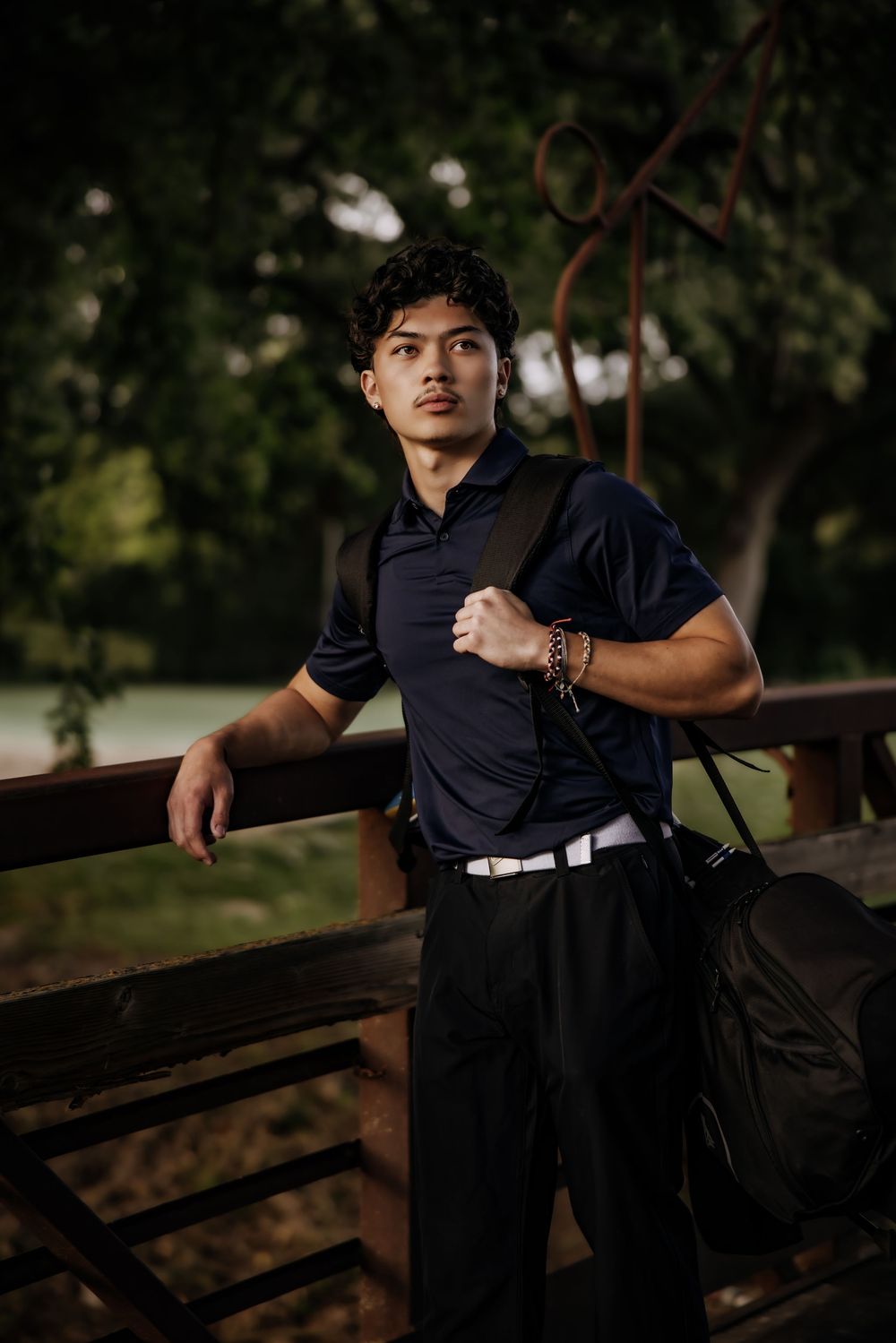 Young man in navy polo shirt carrying a black duffel bag, leaning on a wooden fence in a park.