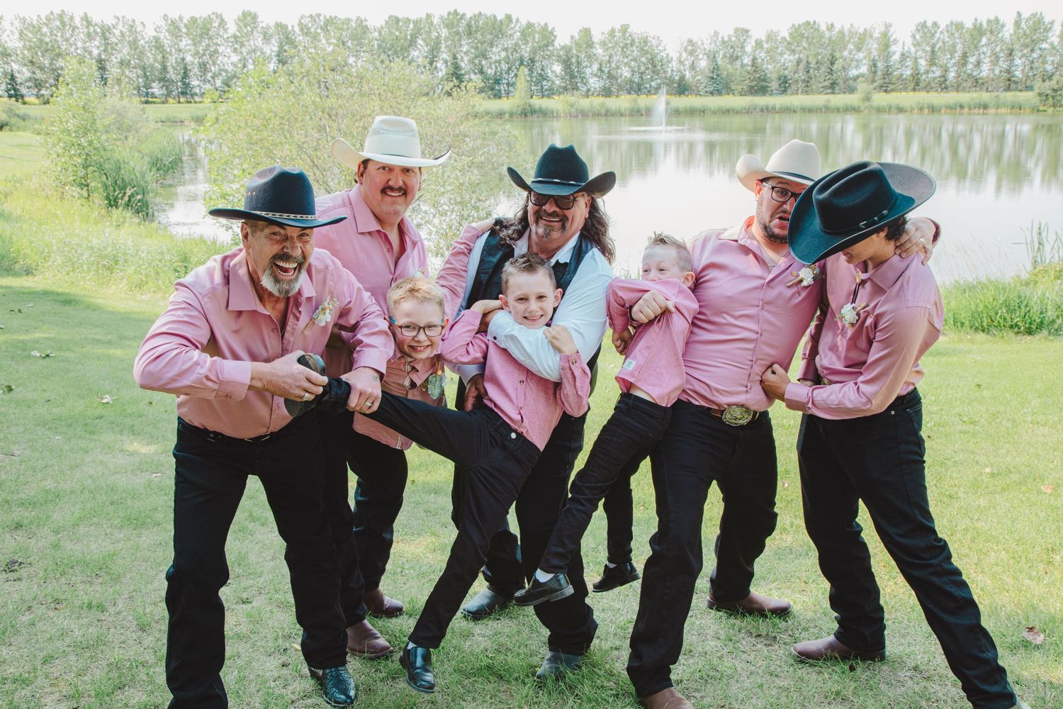 Groomsmen in pink shirts and black cowboy hats playfully lift a child outdoors near a pond at a country wedding.