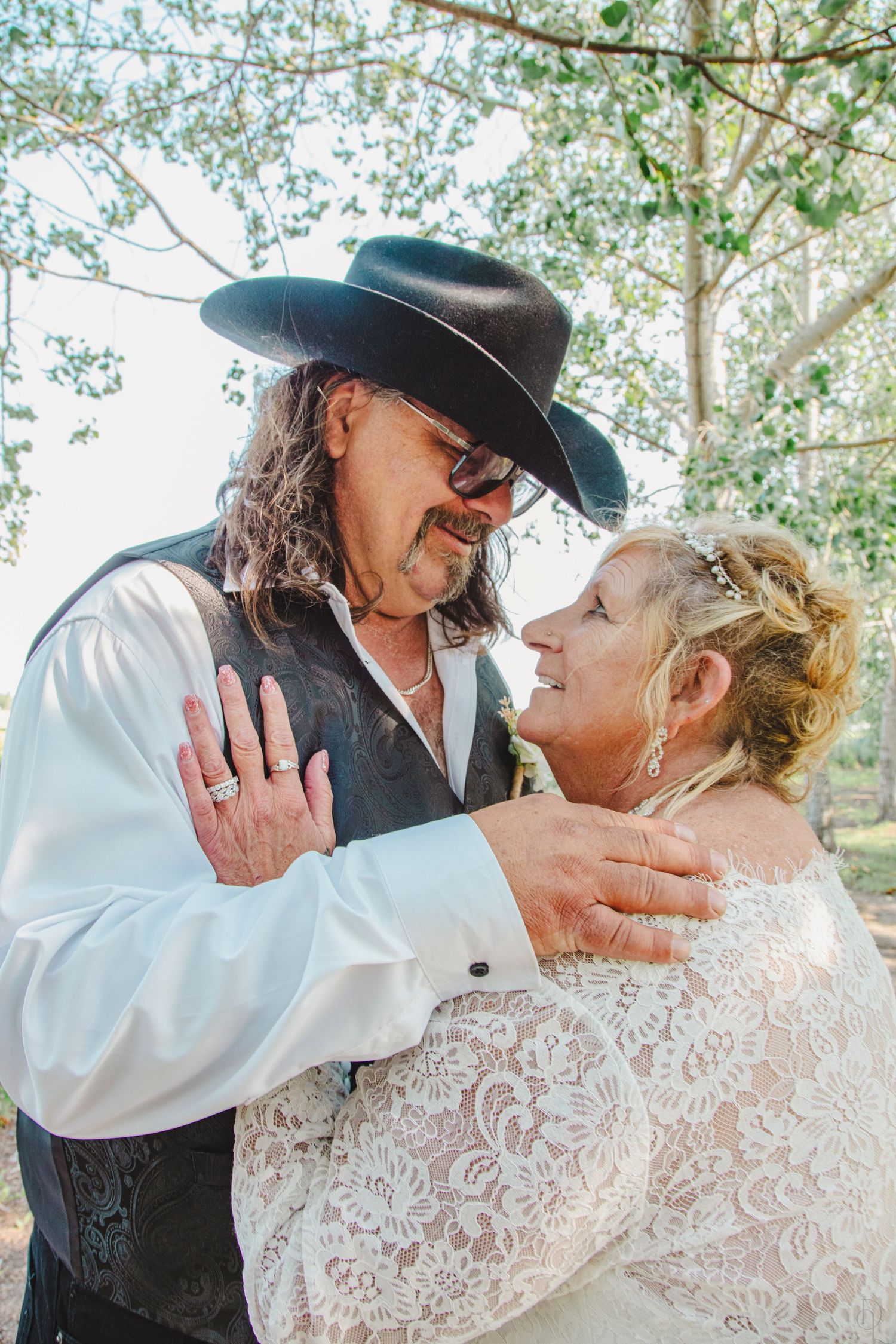 Couple in wedding attire embracing outdoors, groom wearing black cowboy hat and vest, bride in lace dress, smiling at each other.