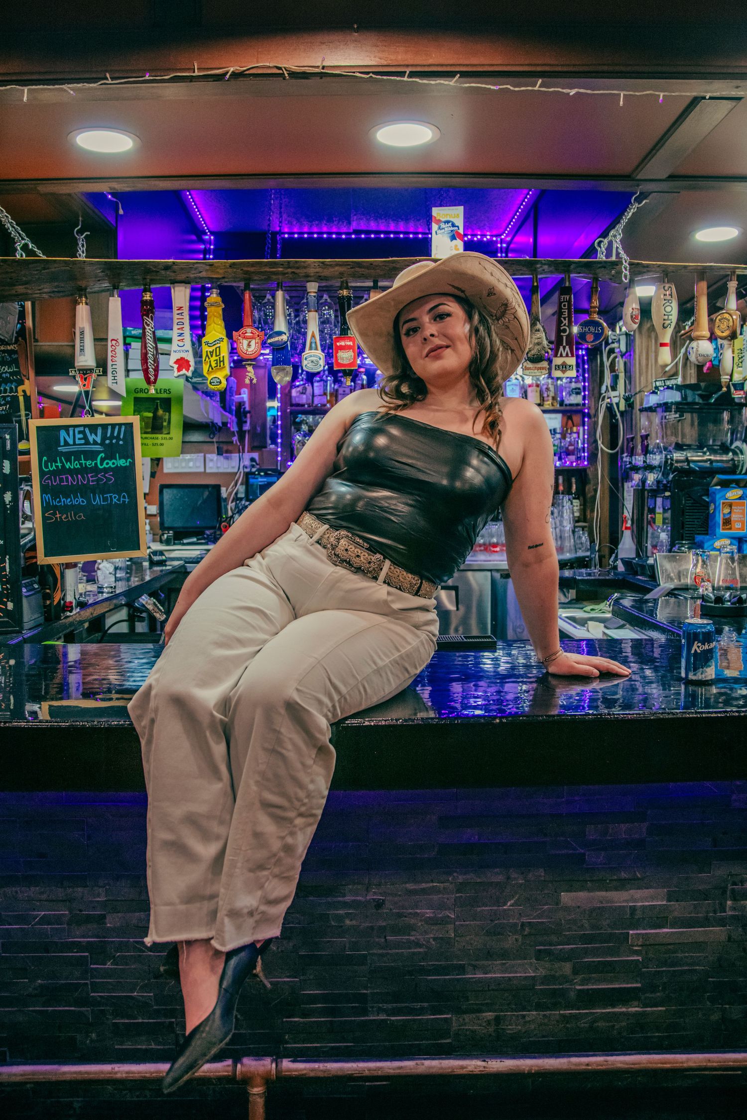 Woman in cowboy hat and black top sits on bar counter in front of colorful lit shelves in a trendy bar.
