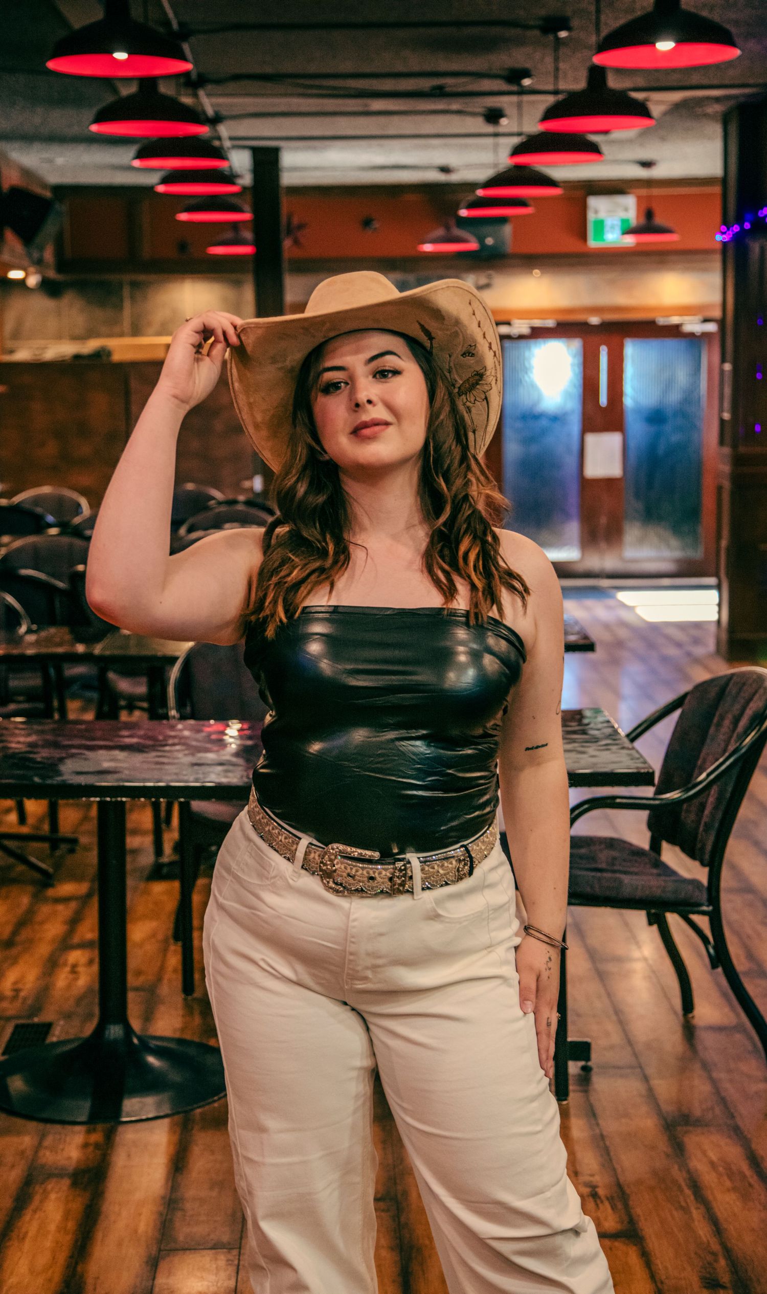 Woman in black leather top, white jeans, and tan cowboy hat posing in a bar with red pendant lights.