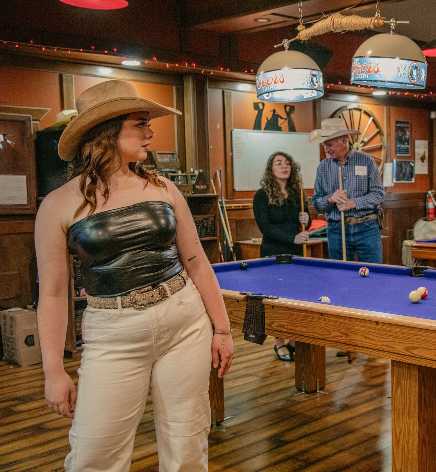 Woman in cowboy hat and leather top stands near a pool table in a rustic bar with guests socializing behind her.