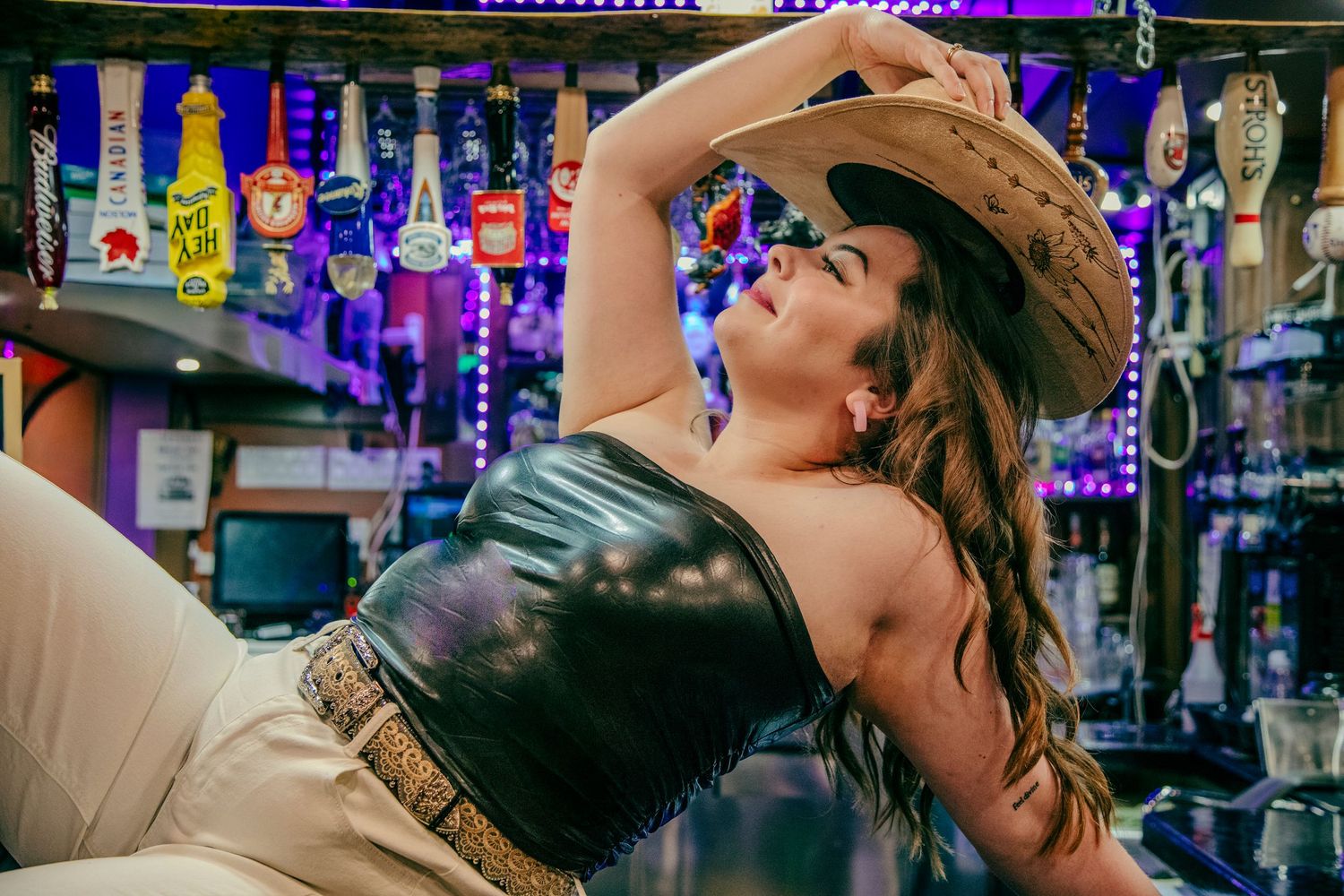 Woman in cowboy hat and black leather top posing at a colorful bar with beer taps in background.