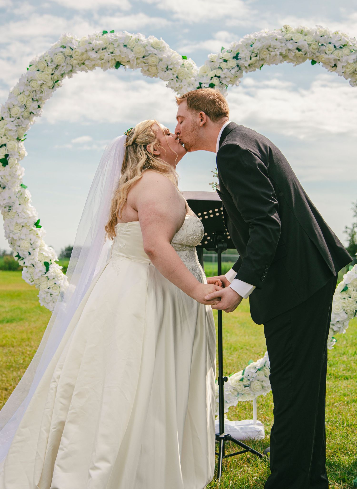 Bride and groom share first kiss under white floral heart arch at outdoor wedding ceremony.