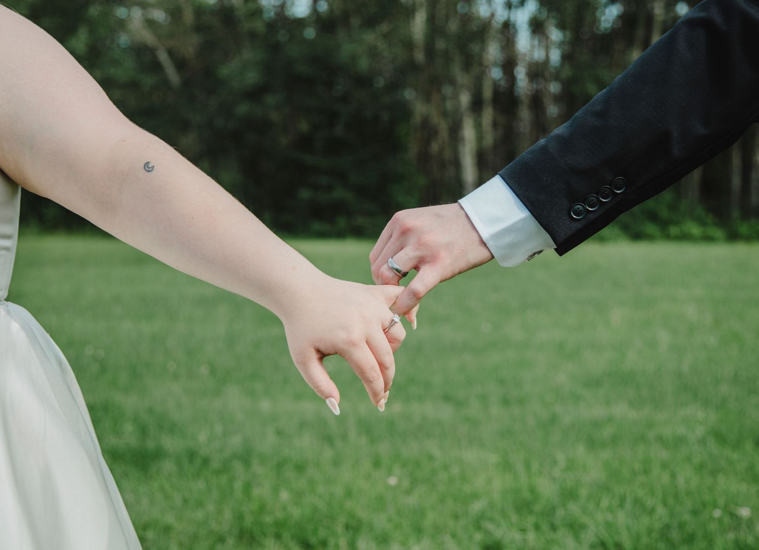Bride and groom holding hands outdoors on green grass, showing wedding rings, in elegant wedding attire.