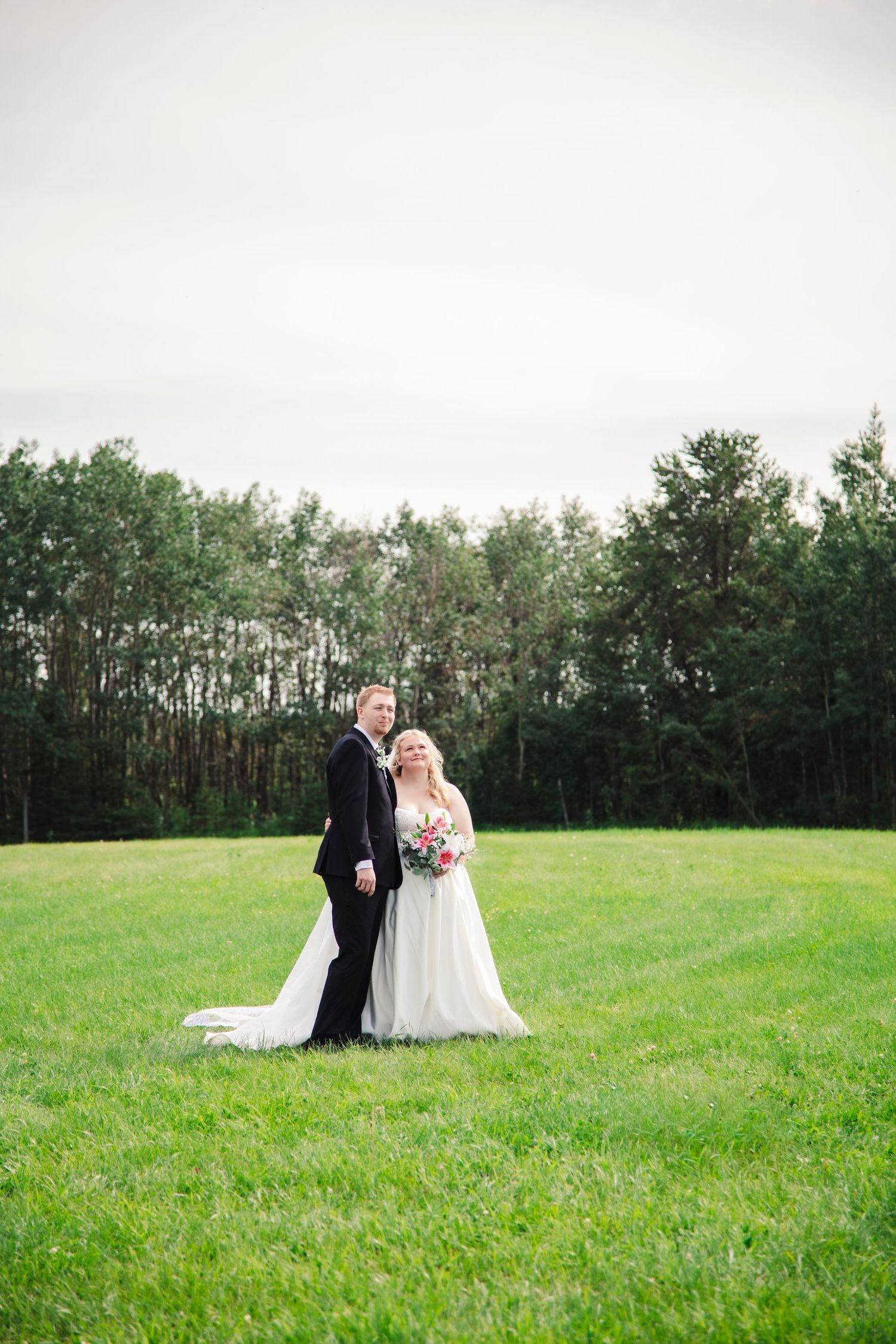 Bride and groom pose on lush green field with forest backdrop on their wedding day.