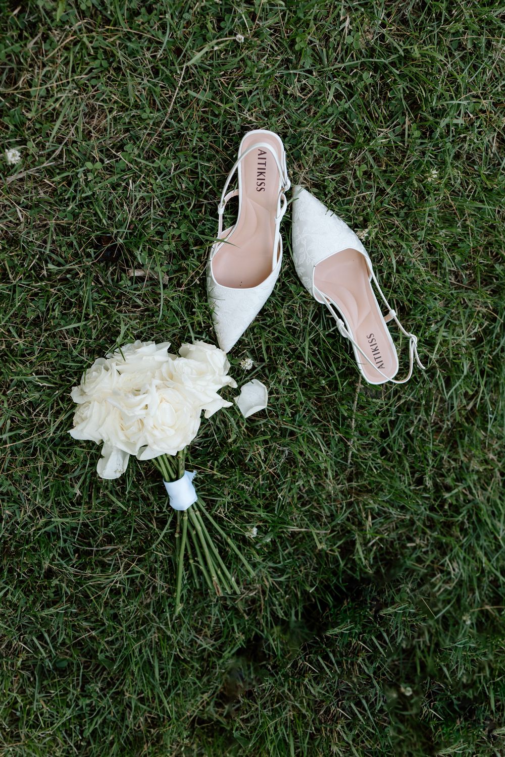 A white floral bouquet and elegant bridal shoes arranged on green grass at different angles.