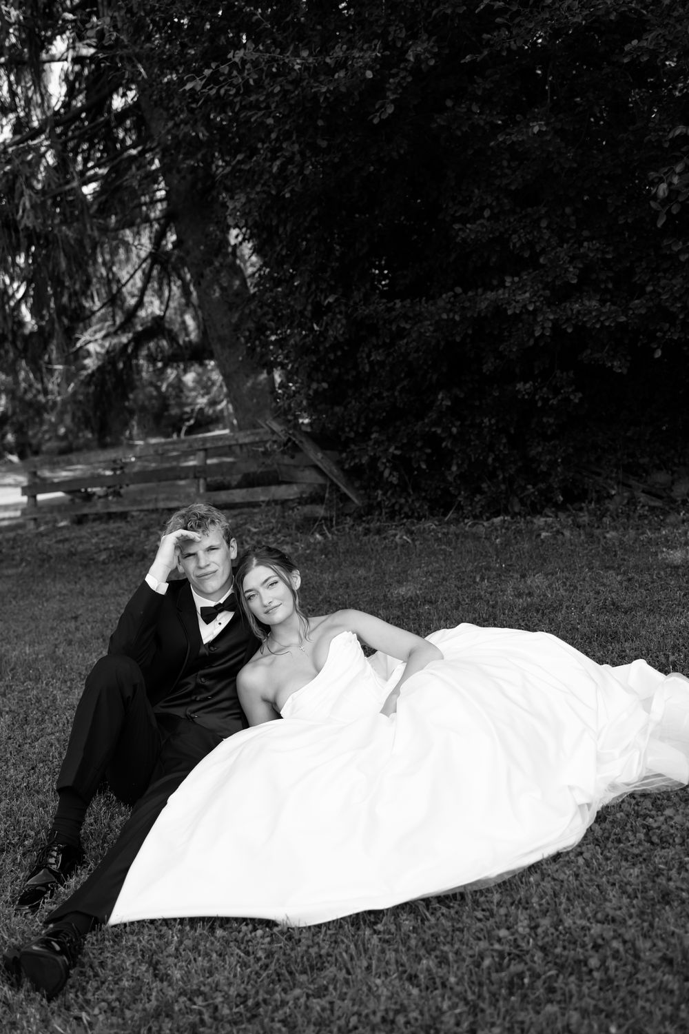 A bride in a white wedding gown and a groom in formal attire relax together on grass in a black and white photo.