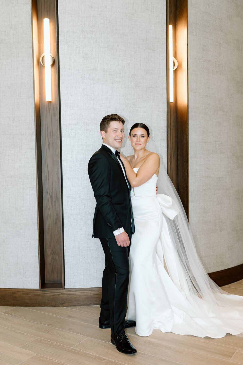 A bride in a white gown and groom in a black suit pose together between two wall sconces.