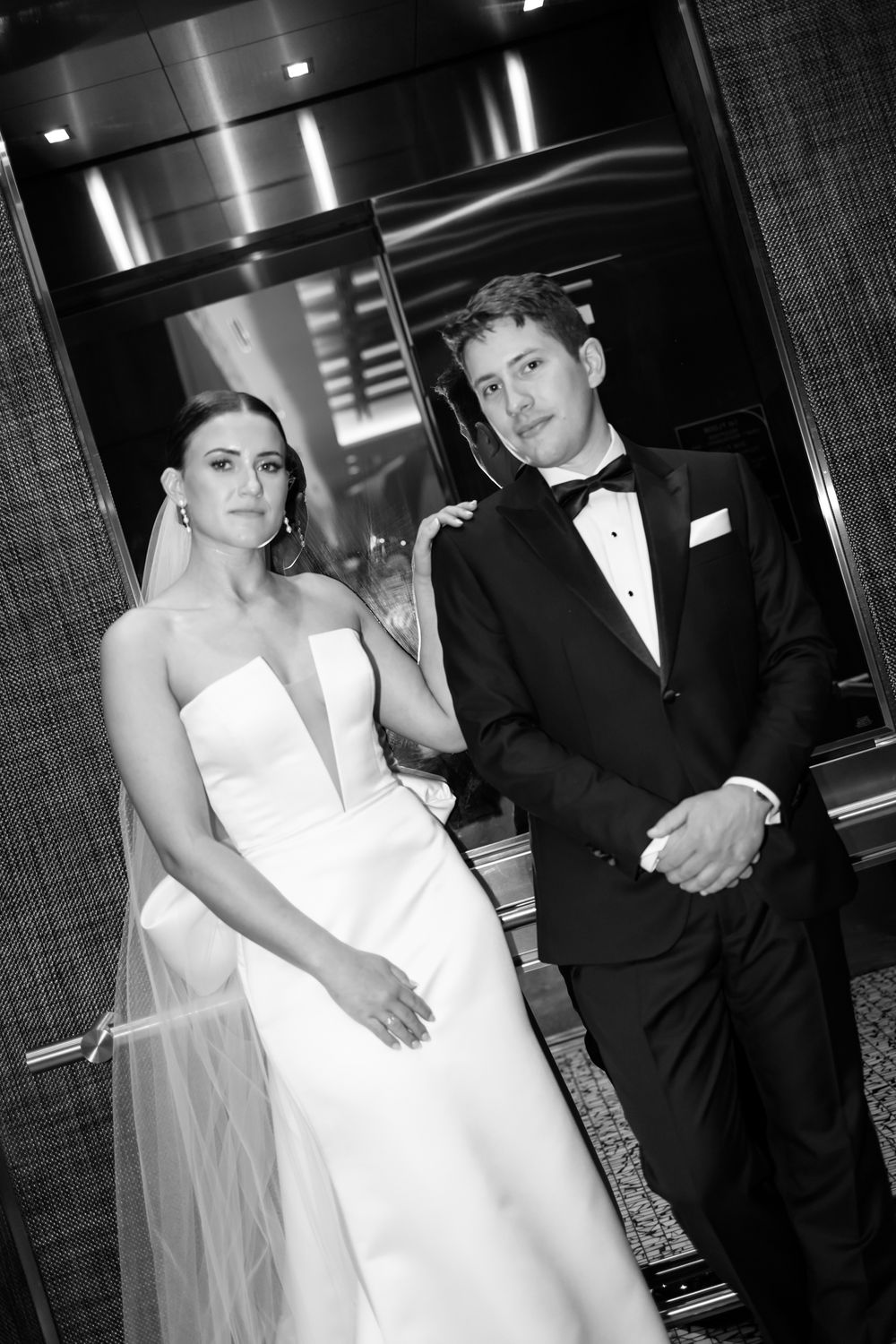 Black and white wedding photo of a bride in a strapless gown and groom in tuxedo posed elegantly in a modern hallway.