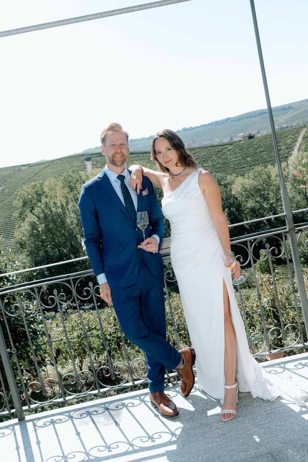 A couple in wedding attire share a candid moment on a balcony overlooking scenic countryside.