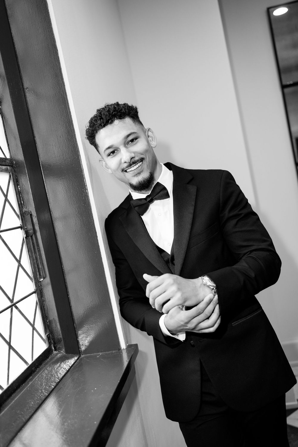 Smiling groom in black tuxedo and bow tie leaning against wall by window in black and white portrait.