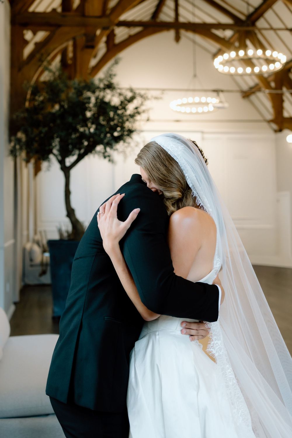 Bride and groom share an emotional embrace in an elegant white wedding venue with wooden beams and chandelier.