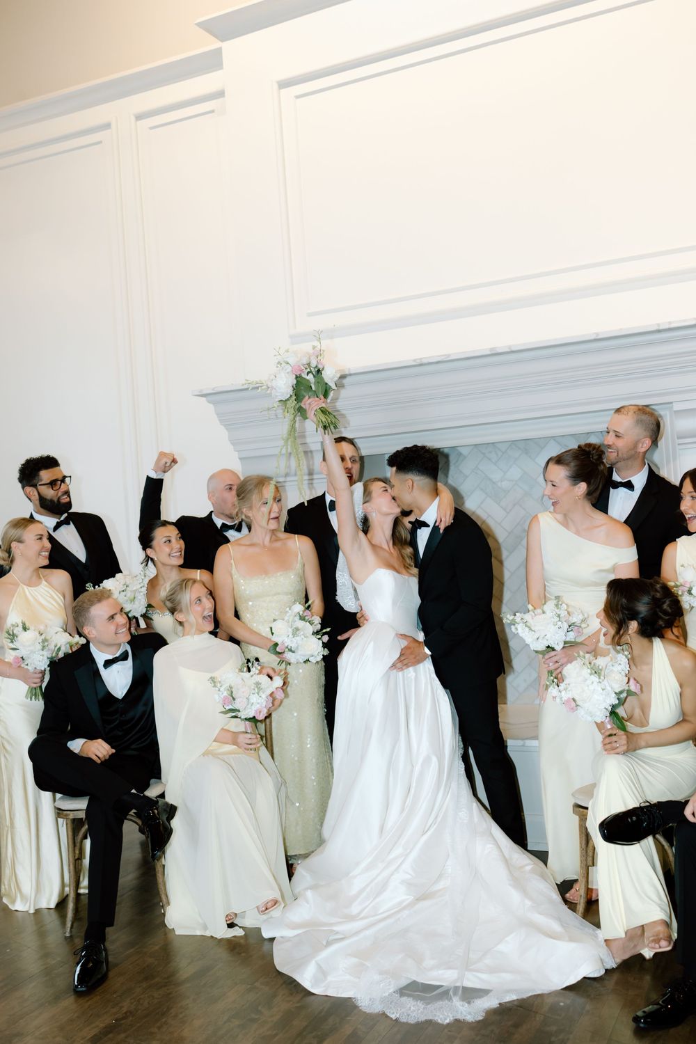 Wedding party poses near fireplace, bride and groom kissing while guests celebrate in elegant black and white attire.