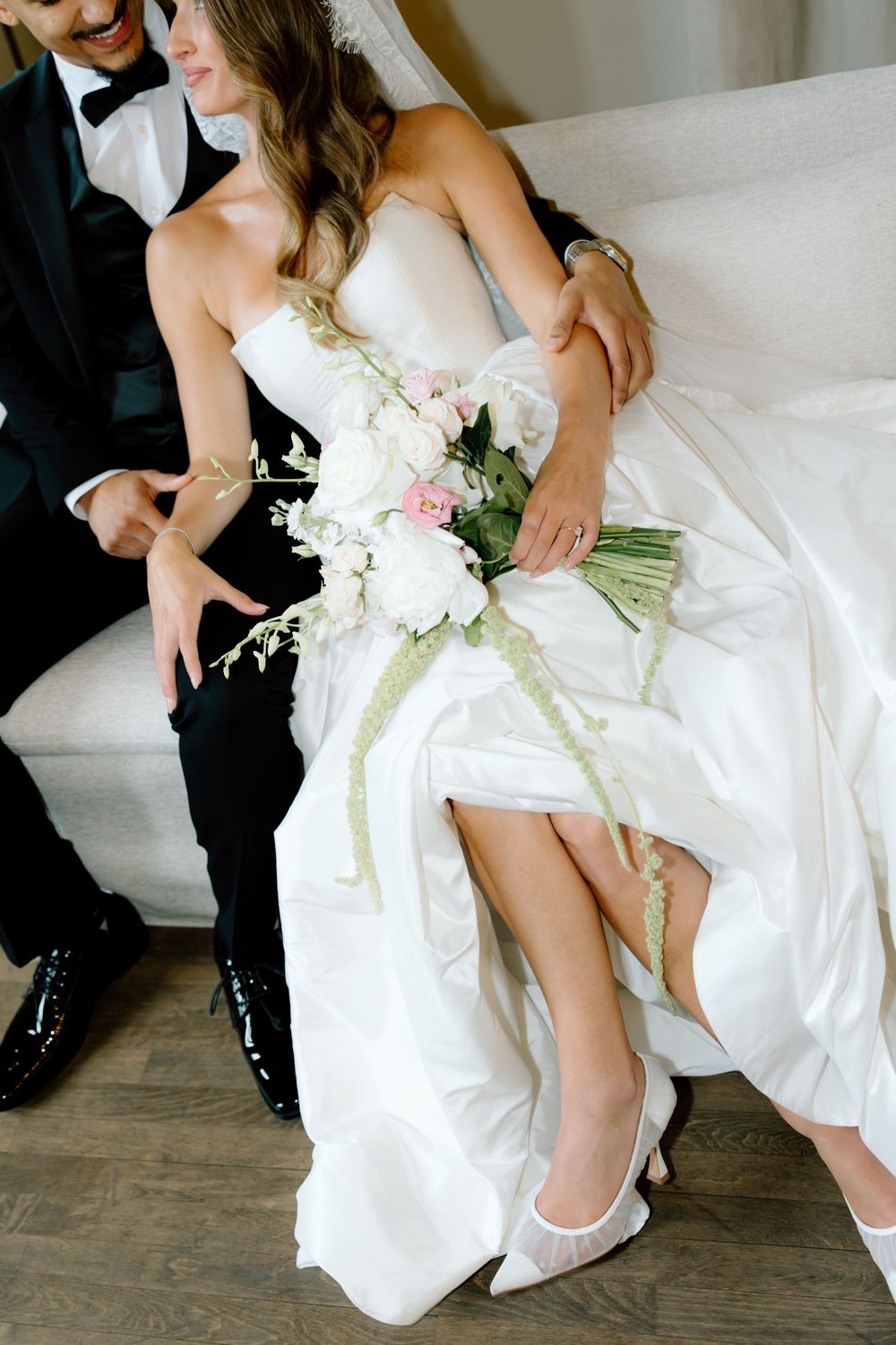 Bride in white strapless gown holding pink floral bouquet sits beside groom in black tuxedo on white sofa.