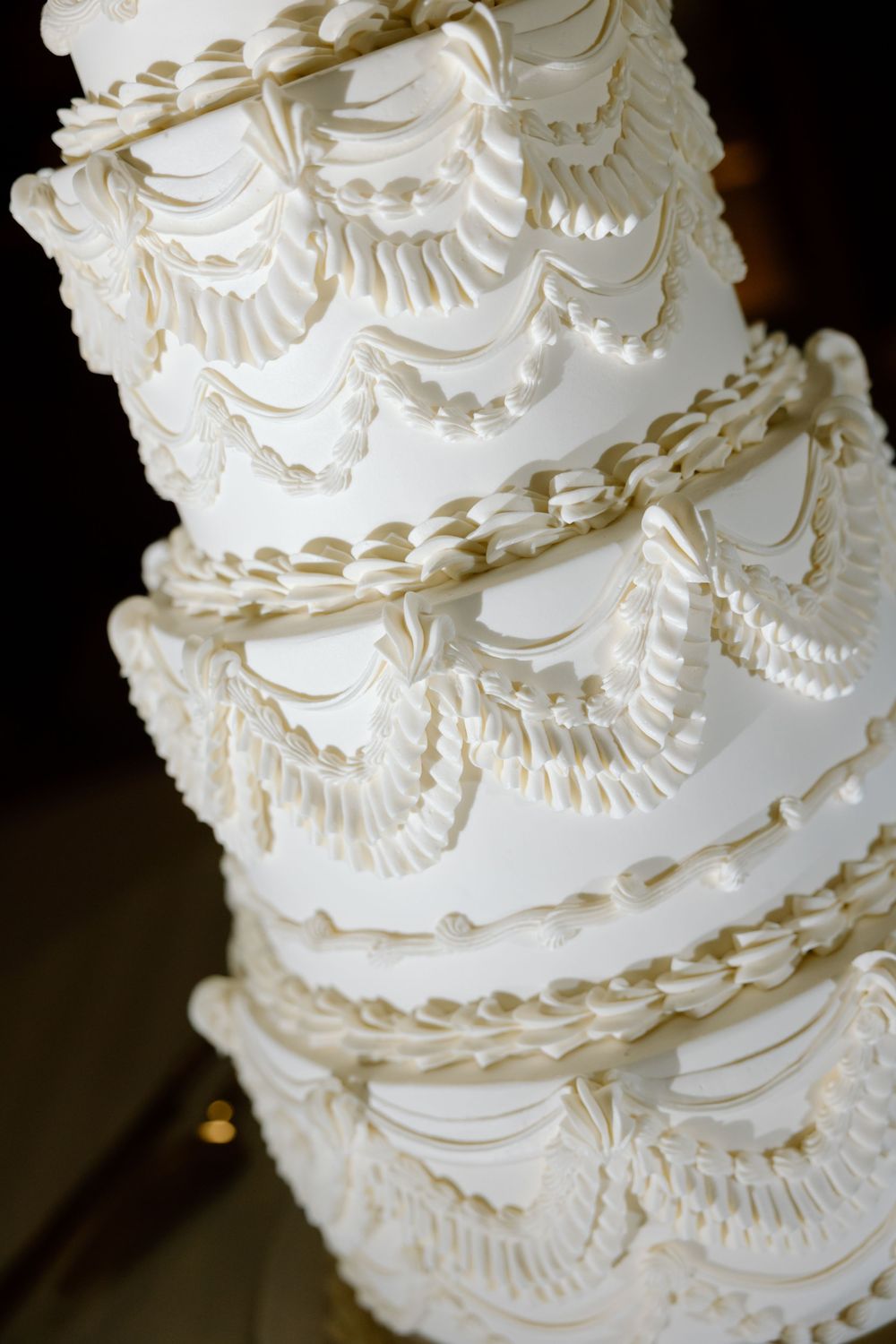 Elegant multi-tiered white wedding cake with intricate fondant draping and decorative piping details on dark background.