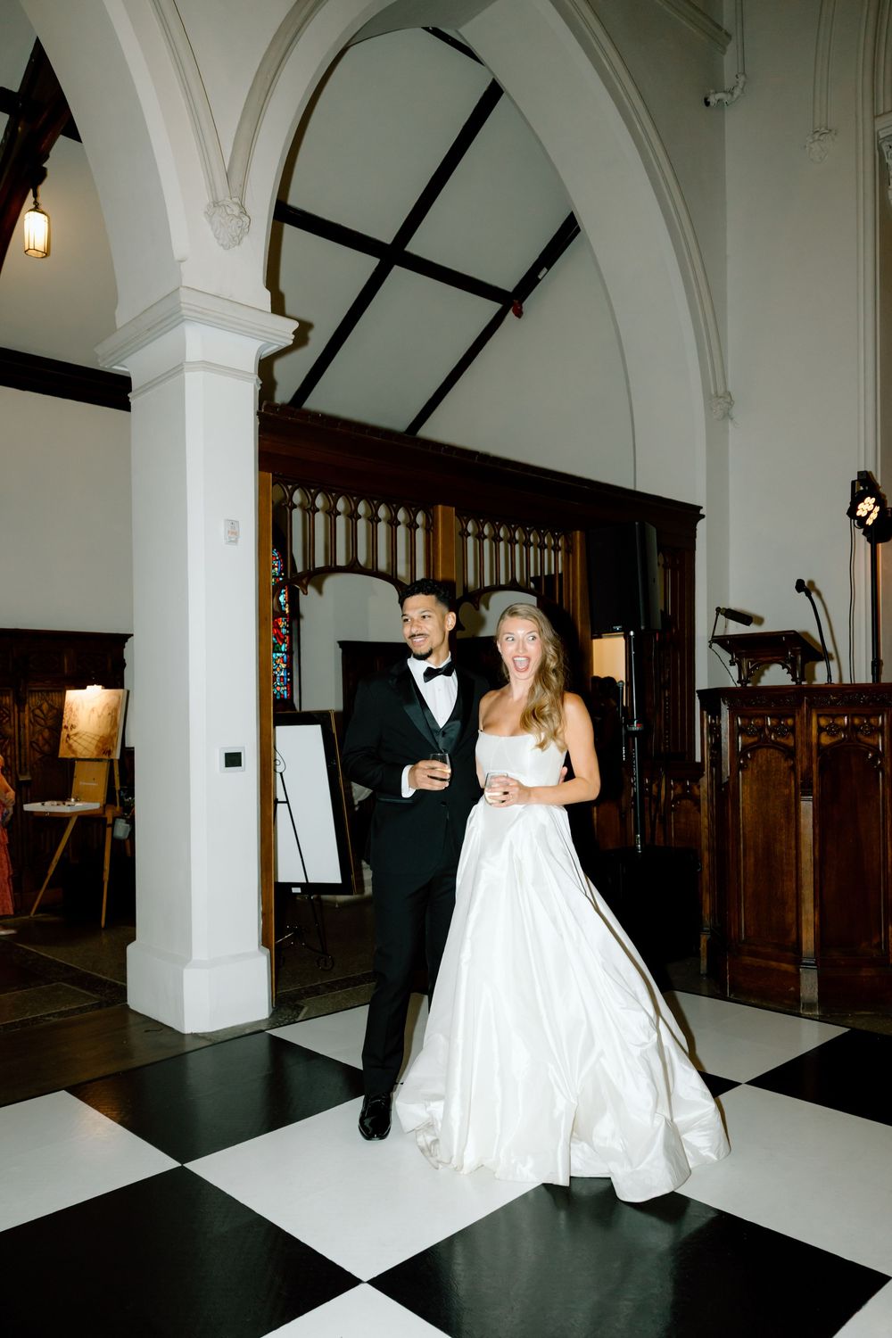 Bride in white ball gown and groom in tuxedo walking on checkered floor in elegant church venue.