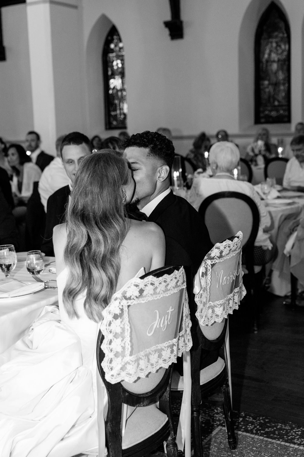 Black and white photo of bride and groom sharing a kiss at their wedding reception table in a church venue.