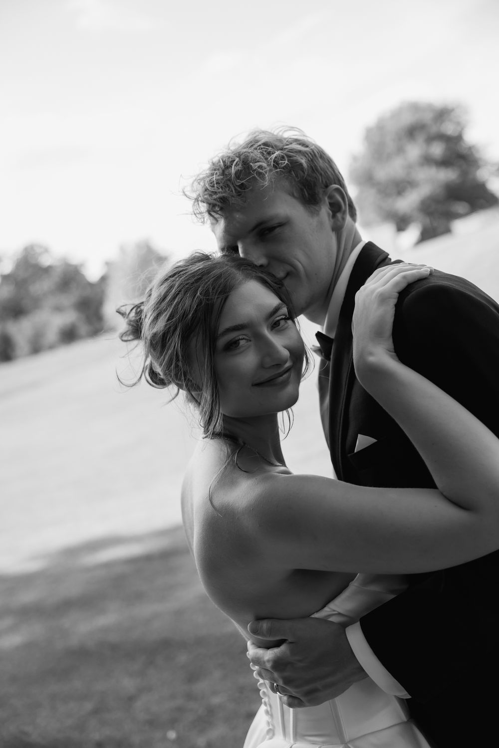 A romantic black and white sequence of strapless-gown bride and suited groom embracing on grassy hillside at sunset.
