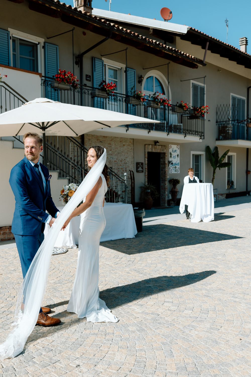 A couple in wedding attire shares a playful moment on a cobblestone patio with white umbrellas and balconies in view.