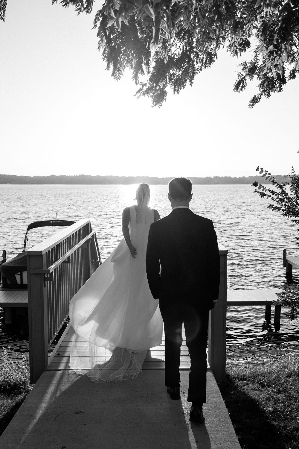 Black and white photo of bride and groom standing on a dock overlooking a sunlit lake on their wedding day.