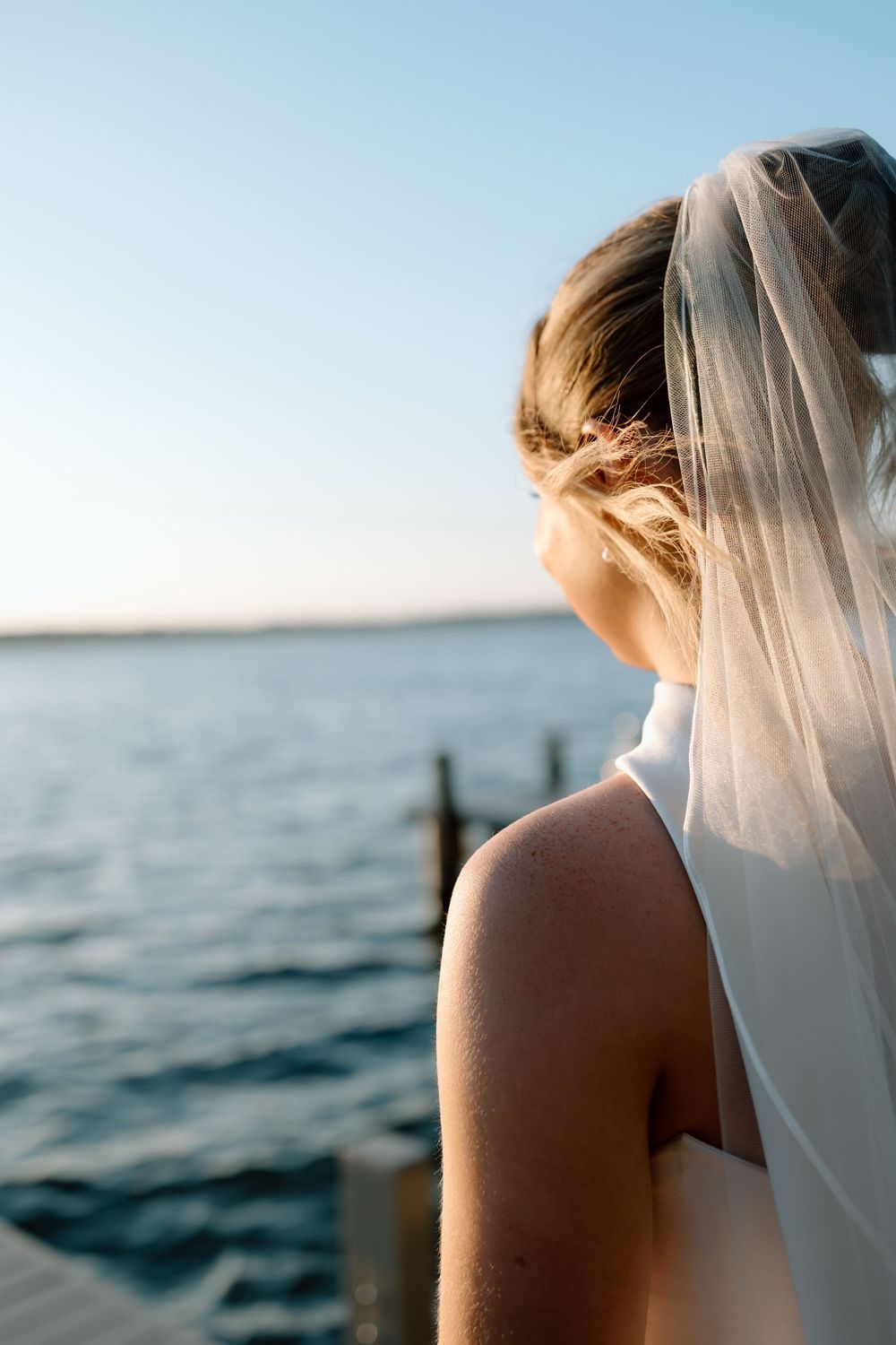 Bride in white veil gazing at calm ocean water from a dock during golden hour sunset.